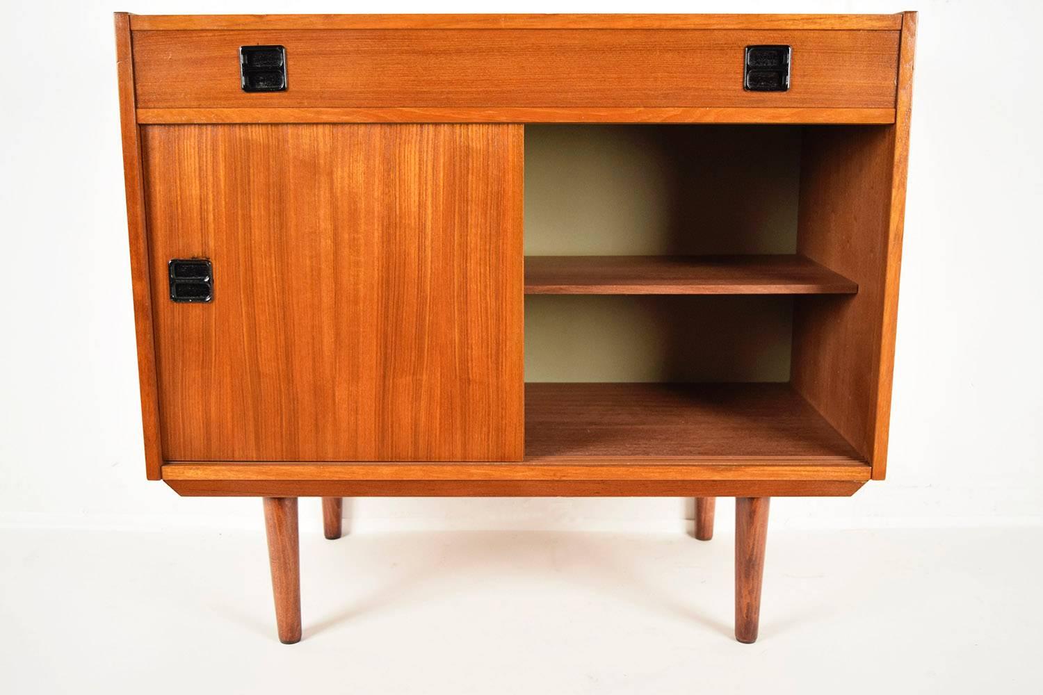 This is a 1960s Danish cabinet. Teakwood, with original Provincial color finish. Features a wooden top, followed by one large front drawer with two black color handles. Bottom has two sliding doors with black color handles and one wooden shelve