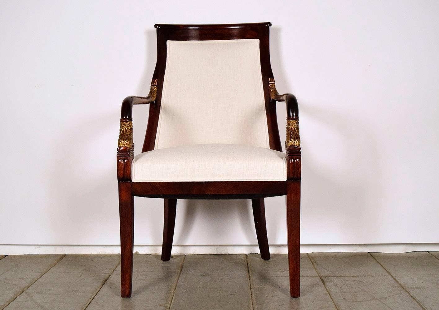 This set of eight Empire-style dining chairs feature frames made from mahogany wood that has been stained in a deep and rich color stain. The frames feature a curved back that leads into beautiful carved arms with gilded leaf motif accents. The