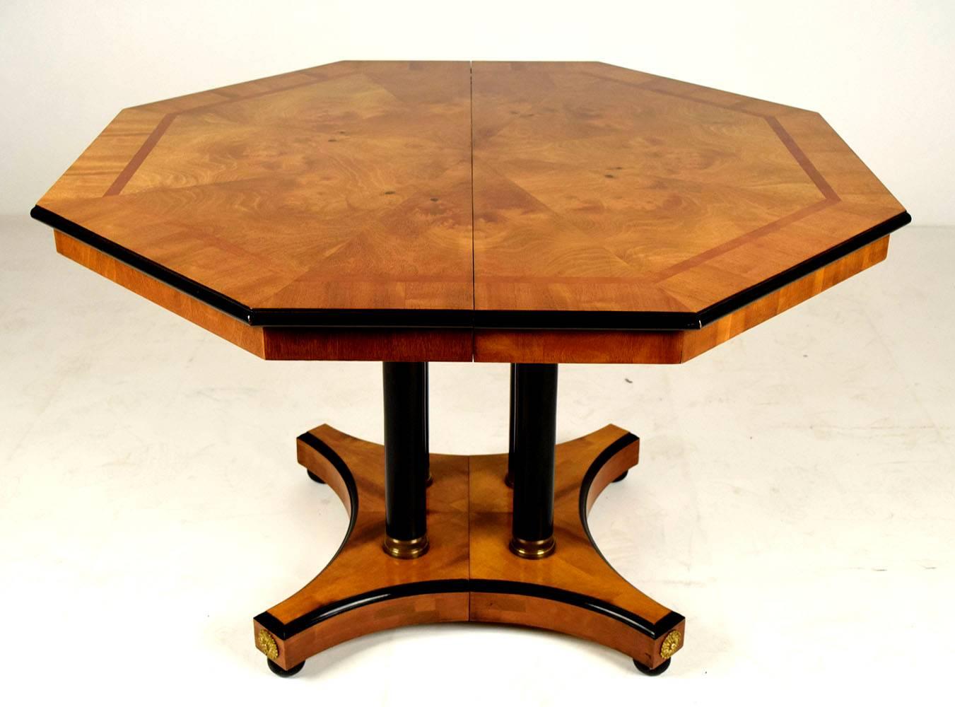 This beautiful octagonal shaped dining table is made of solid wood and covered in outstanding burl veneers with a golden yellow finish and black accent on the top sides. The elegant stretched legs have four black accent columns and brass rings at