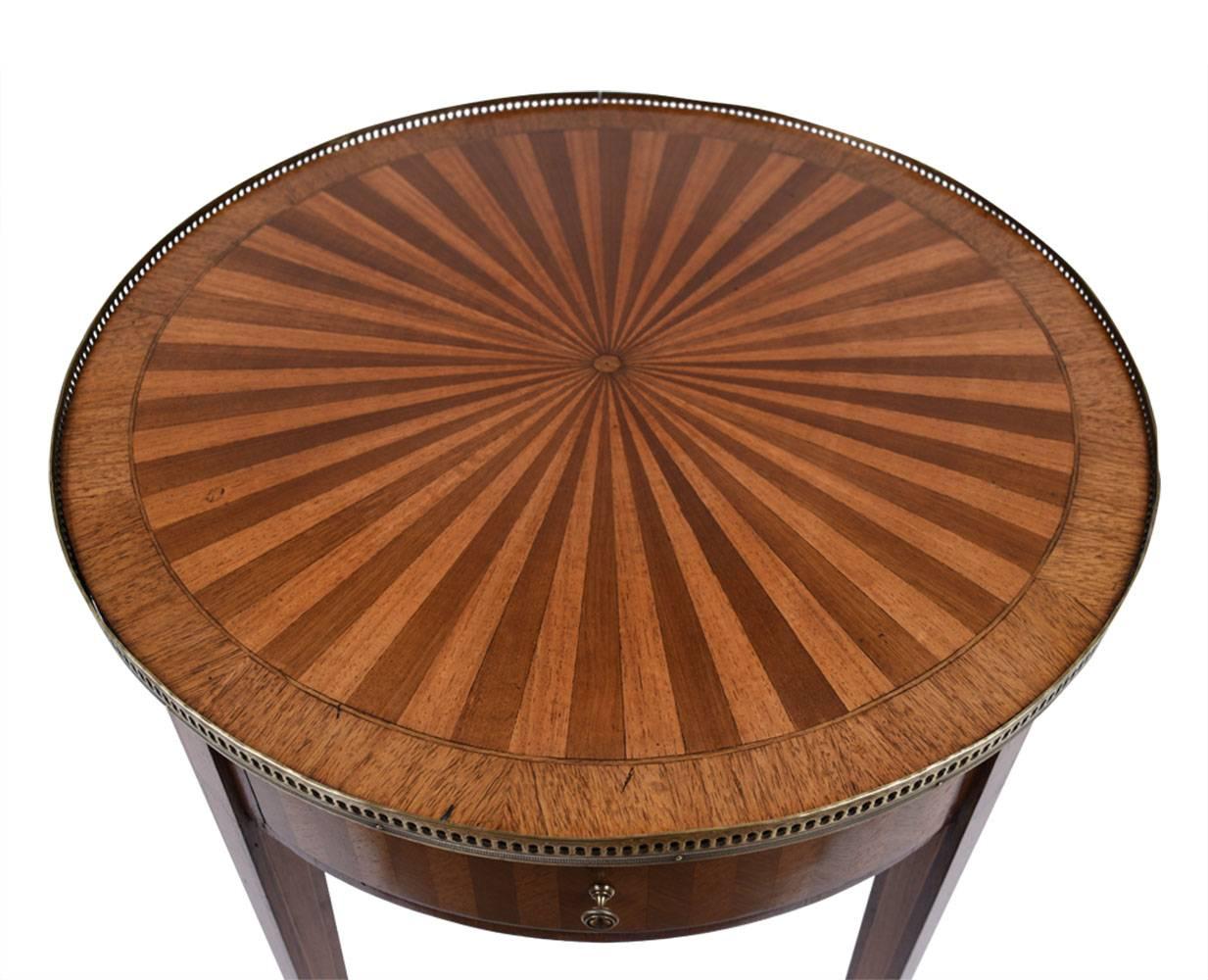This 1900s Antique French Louis XVI-style parlor table is made of walnut wood finished in a walnut color stain. The side tables are adorned with inlaid designs. On the top the inlaid details are in a sunburst pattern that is accentuated by the brass