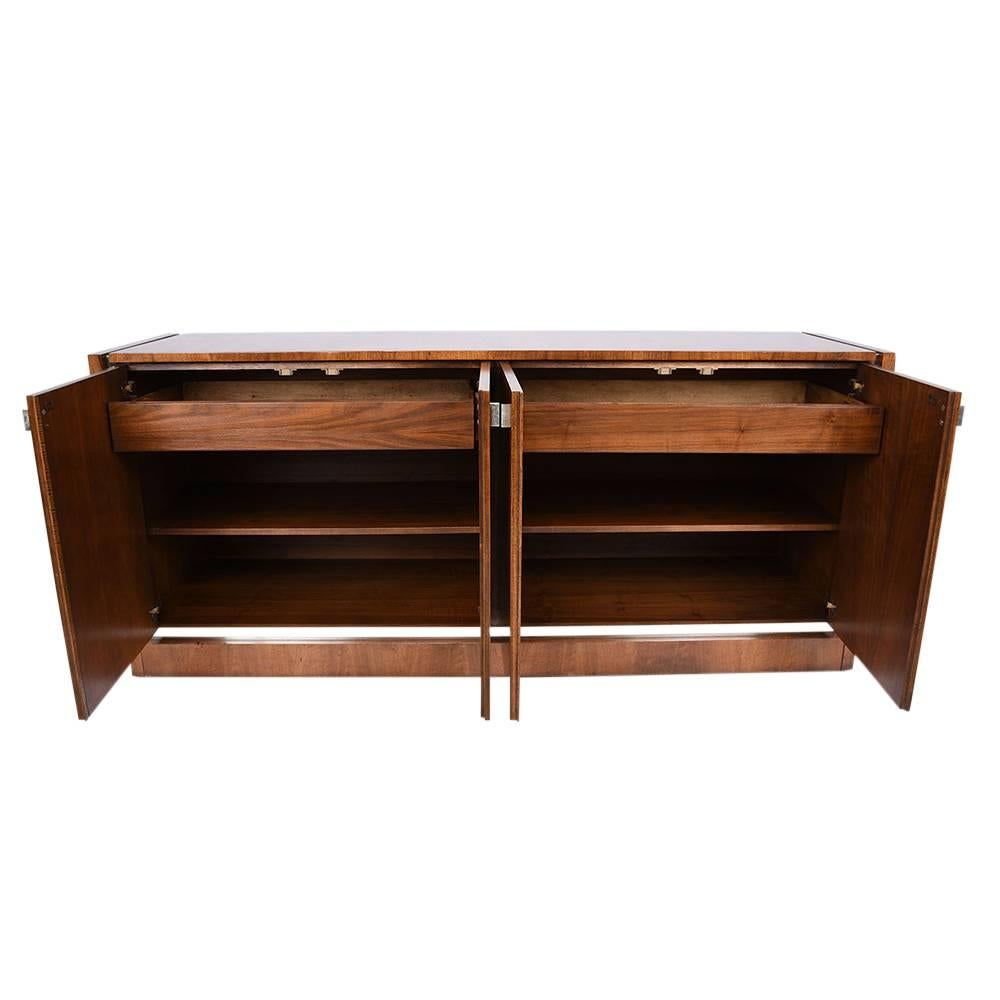 This 1970s vintage Mid-Century Modern style credenza is designed by Founders. The credenza is made of walnut wood that has been stained in a beautiful medium wood tone color. The wood top is accented by chrome molding along the sides and down the