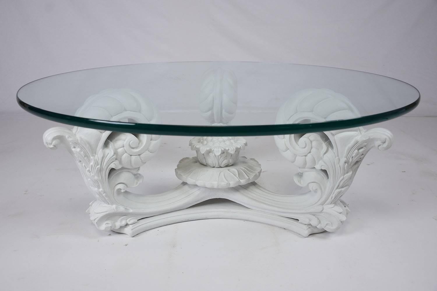 This 1960s Hollywood Regency-style coffee table features an ornately carved wood coffee table that has been painted in a pure white satin finish. The scroll arm designed base features acanthus leave decorations with a beautiful flower in the center.