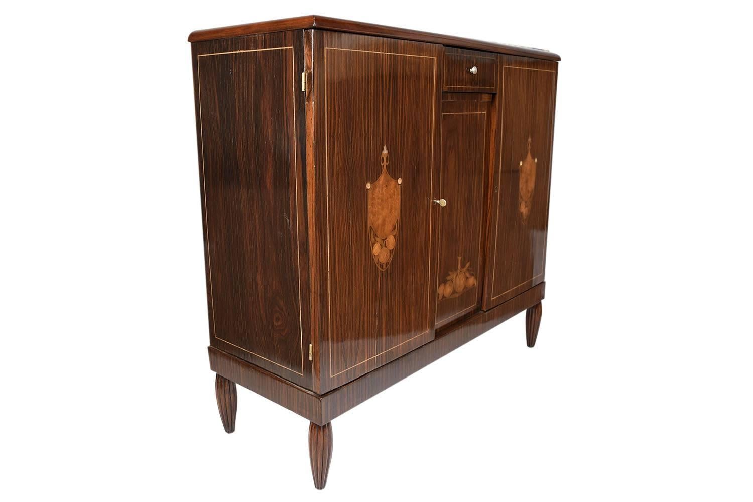 This 1930s Antique French Art Deco-style buffet is made of mahogany wood with beautiful Macassar wood veneers with a lacquered finish. The top features a white marble slab. The facade of the cabinet has inlaid details of two crests with fruit
