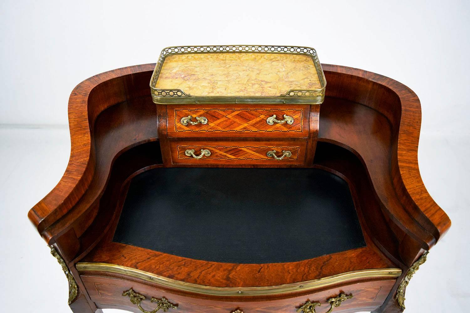 This 1900s antique French Louis XV style desk is made of wood finished in a mahogany color stain. The facade of the desk is adorned with geometric marquetry decorations. Along the top of the desk is a brass gallery with a marble surface. On the top