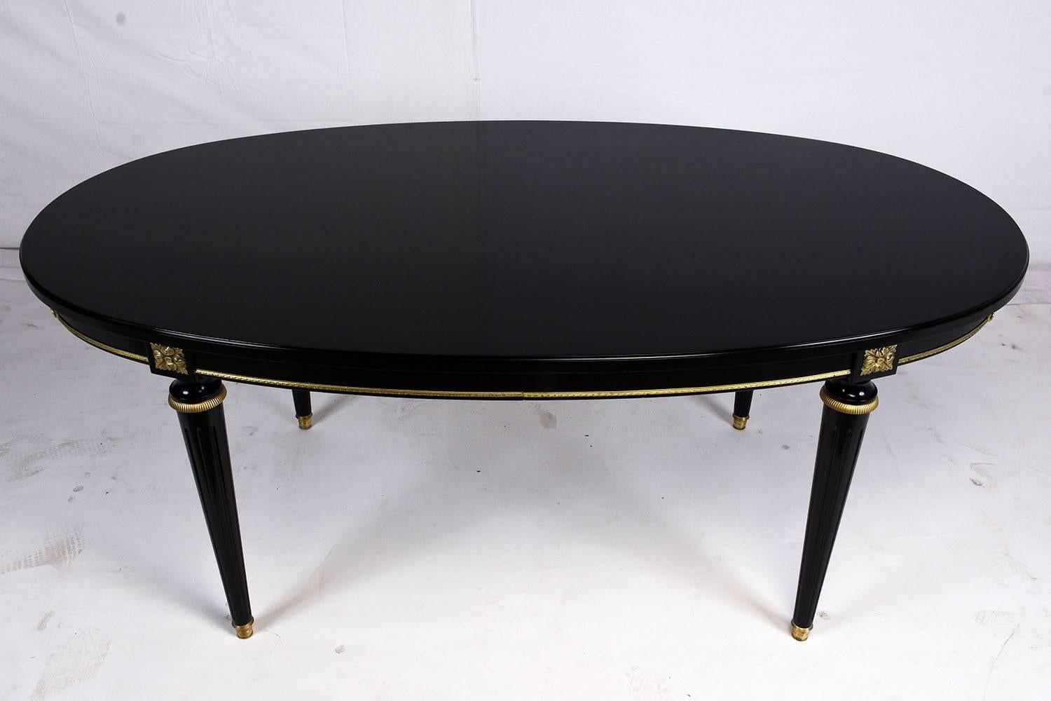 This 1960s French Louis XVI-style dining room table is made of mahogany wood with a beautiful ebonized finish. The oval table is accented by carved wood details of rosettes and beaded bands that have been finished in a gold color. The turned legs