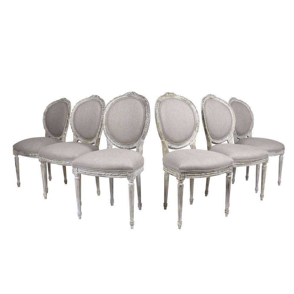This 1900s antique set of eight French Louis XVI-style dining chairs feature carved wood frames that have been painted in a grey and off-white color combination. The oval seat backs features carved garland decorations and have upholstered centers.
