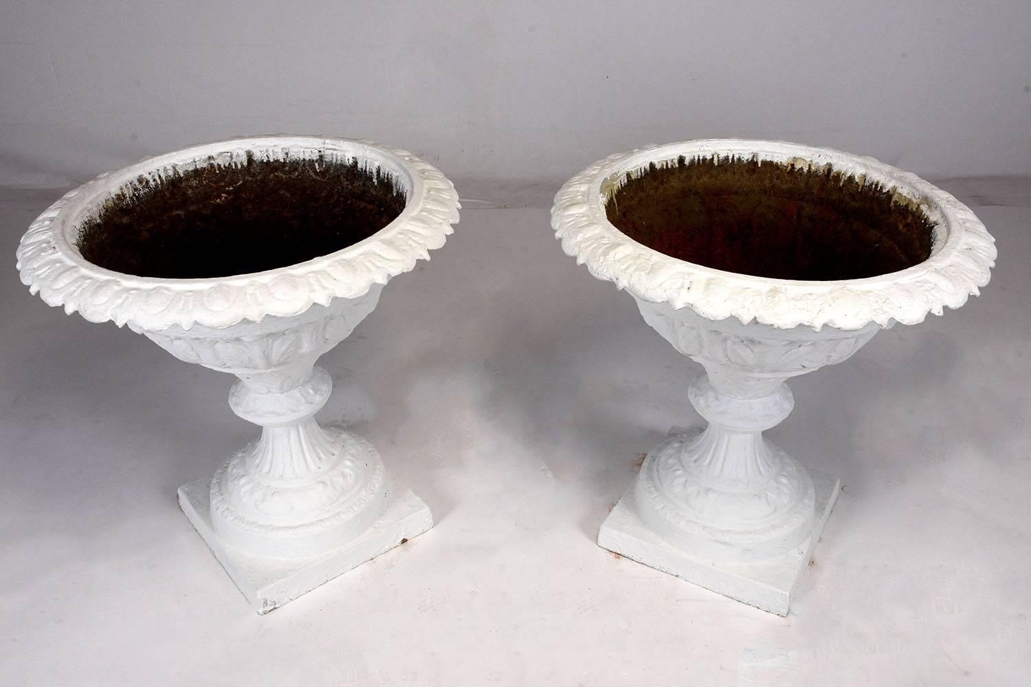This pair of large planters is made from cast iron that has been painted in an off-white color. The planters are beautifully adorned with floral motif designs on the lip, egg and dart decorative band, fleur-de-lis designs, and architectural