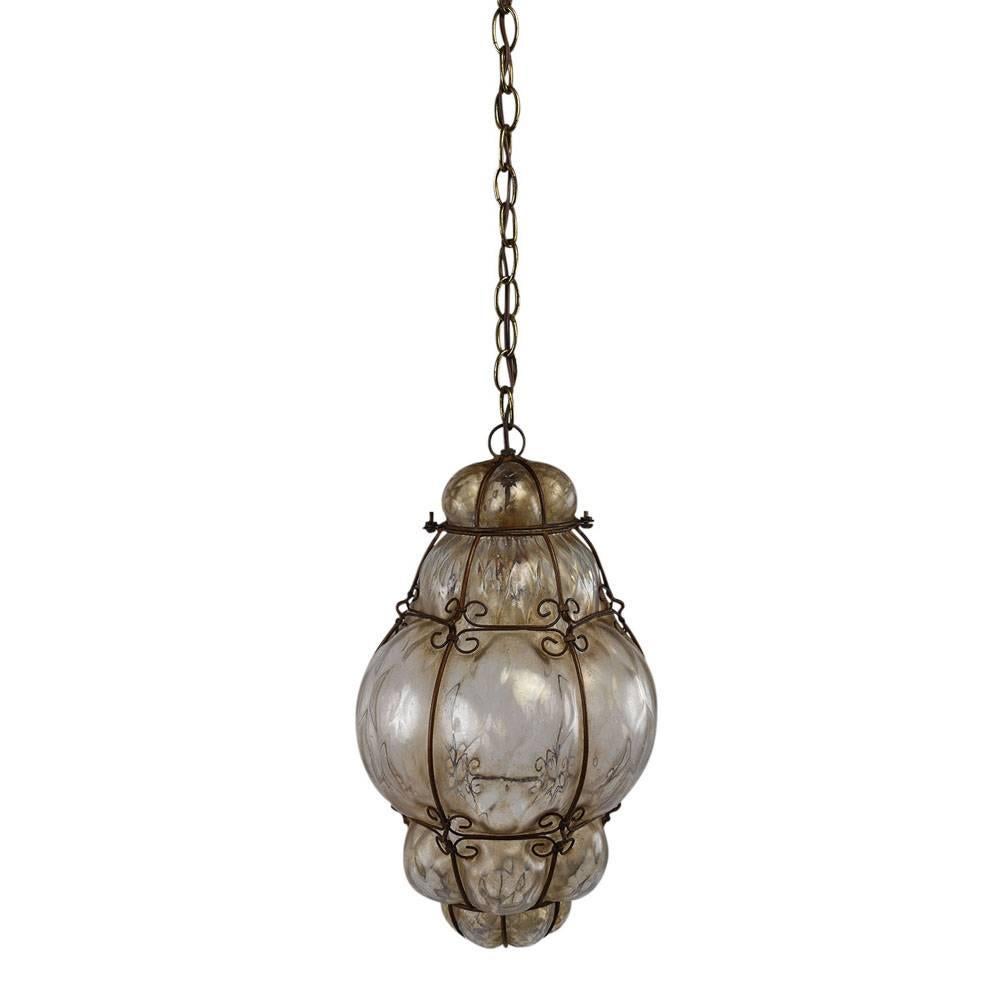 This vintage pendant chandelier is made of blown Murano glass. The bulbous shape of the glass is accented by a delicate metal cage with scroll details. The chandelier features a long chain for easy adjustment. There is one interior light that allows