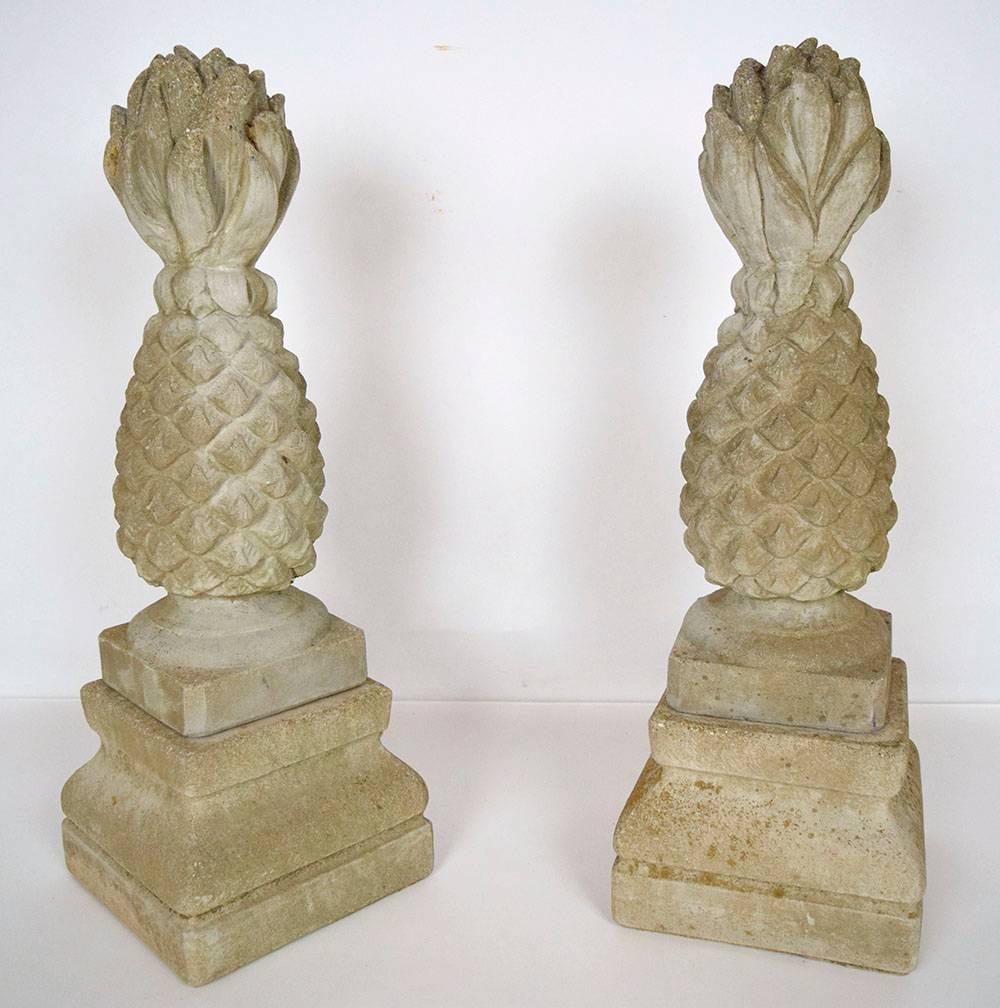 This pair of garden urns is made from cement and has a green coloring added. The urns come in two pieces, the top and the base. The top features a statue of a pineapple in an upright position. The base is designed using architectural motifs.