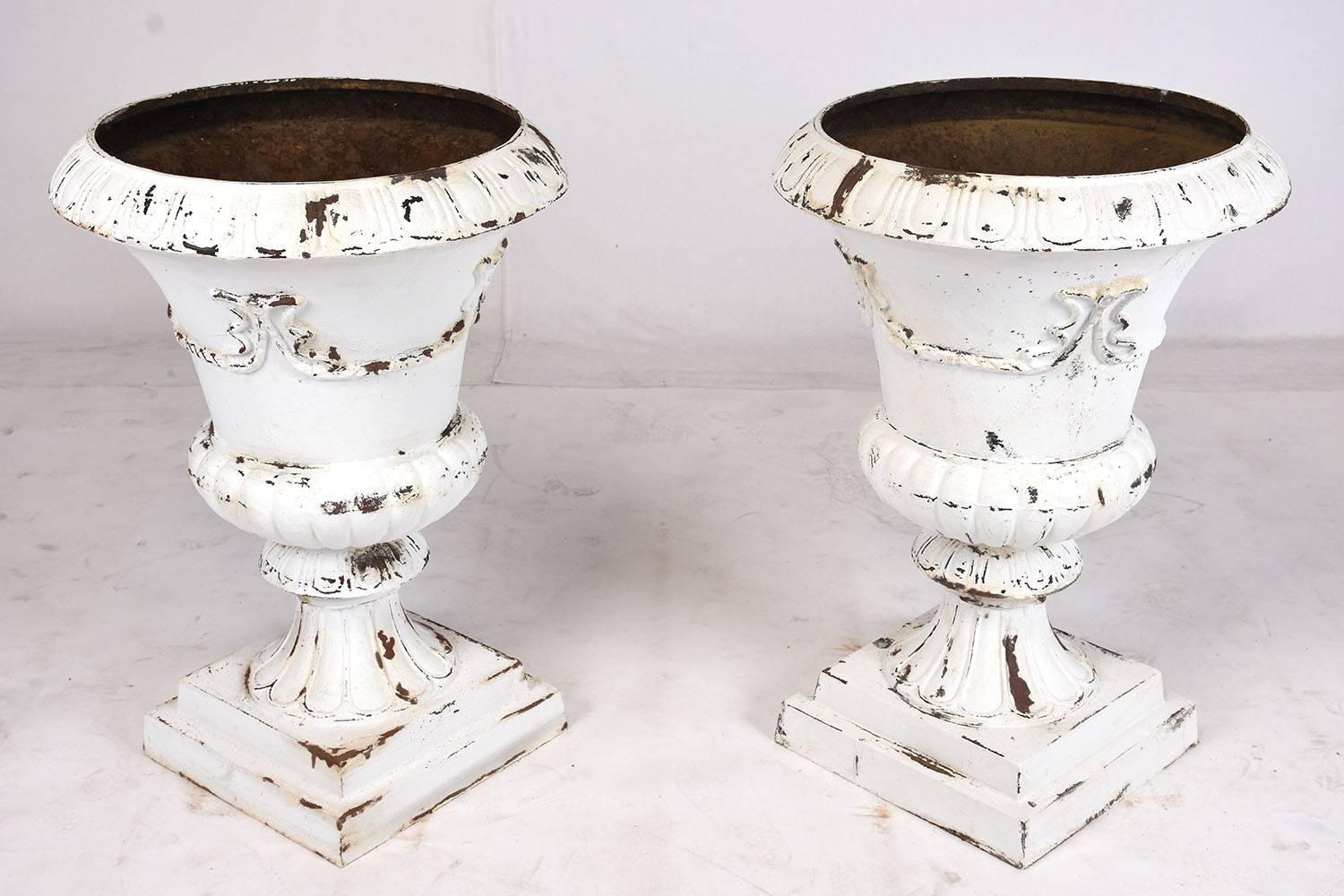 This pair of 1900s antique French neoclassical-style garden urns are made from iron that has been painted white with a distressed finish. There are architectural elements around the urns of egg drop bands and acanthus leave garlands. This pair of