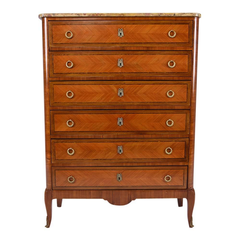 This 1900s antique French Louis XVI-style chest of drawers is made from mahogany wood that has been finished in a medium wood tone stain. The facade of the six drawers feature inlaid details in a herringbone design and two decorative brass round