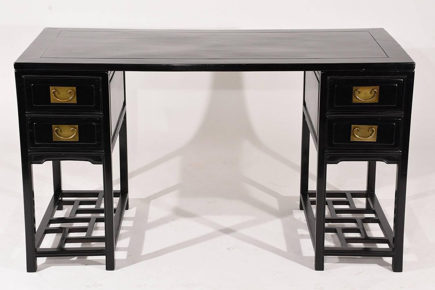This 1970s vintage chinoiserie-style desk is made of wood that has been ebonized with a lacquered finish. The pedestal legs have two drawers each adorned with large brass drawer pulls and an open shelf below with a geometric design. The desk top has