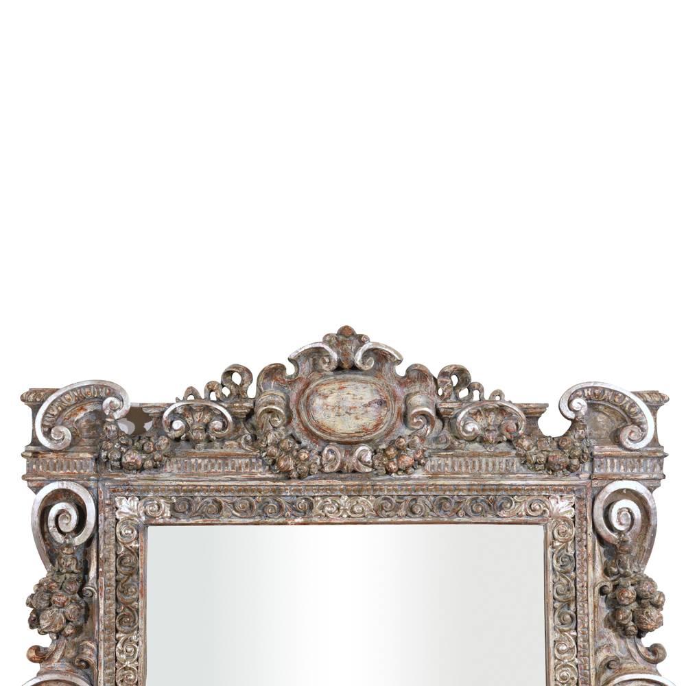 This 1840s antique Baroque-style grand wall mirror features a carved wood frame that has been painted in red, blue, grey, and white color combination with silver leaf accents. The intricately carved frame is adorned with many beautiful details.