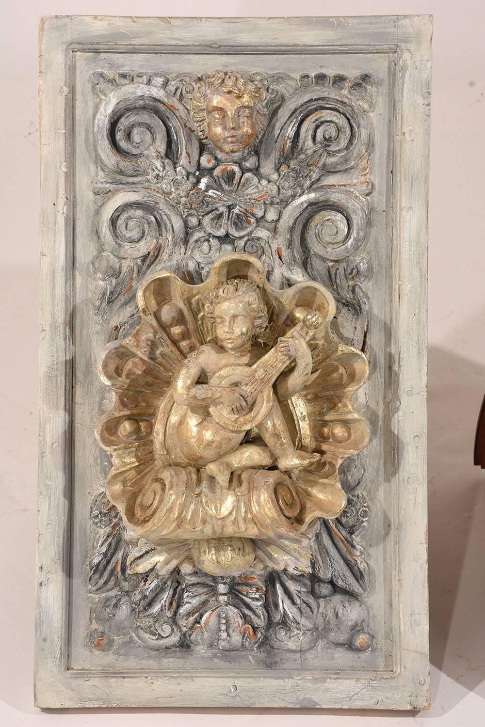 This pair of 1970s Italian Baroque-style wall plaques are made of wood that has been painted in a pale grey, off-white, and gold color combination with a distressed finish. The intricately carved high relief depicts two cherubs playing musical