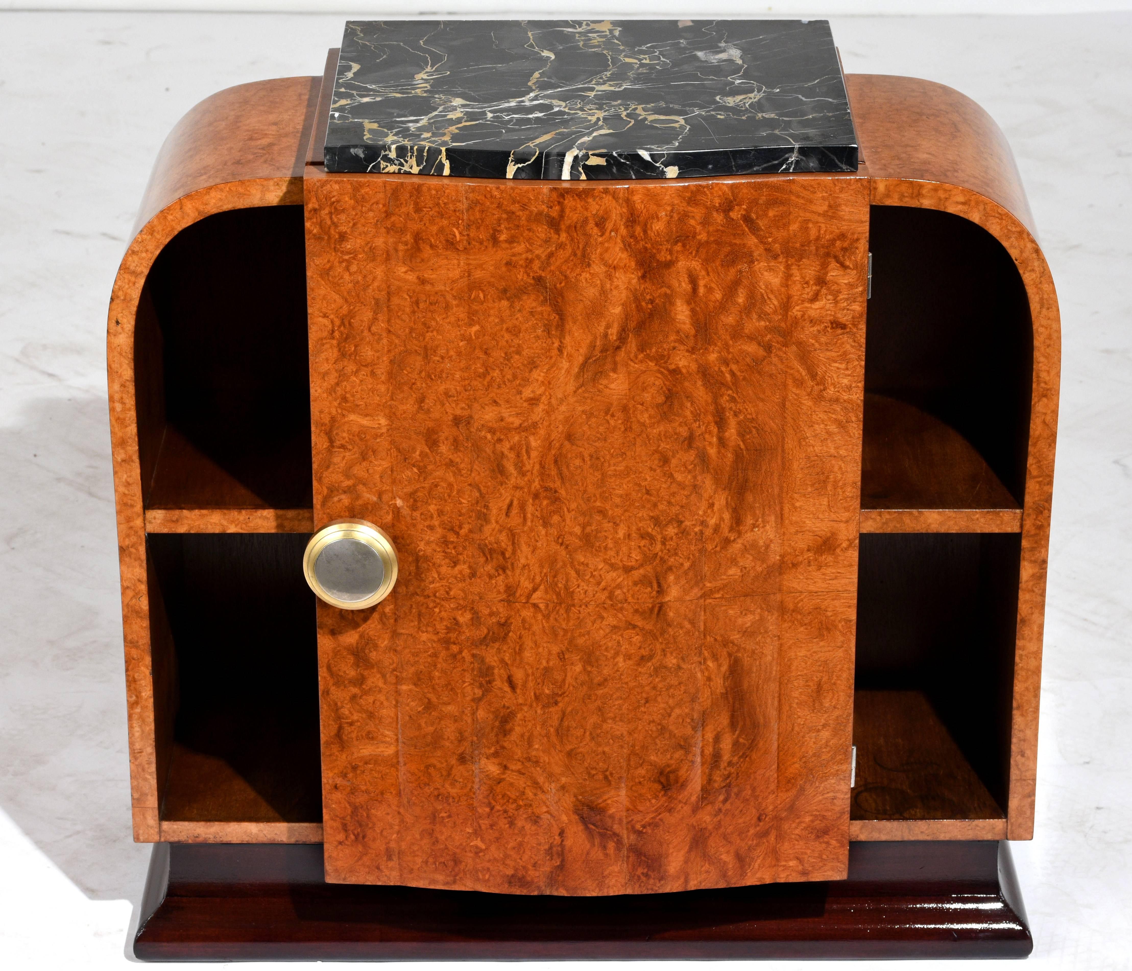 This 1930s French Art Deco-style nightstand is made from wood finished in a walnut color. The front and sides feature sleek lines and curved edges and are adorned with burled walnut wood veneers. The top features a black marble section with white