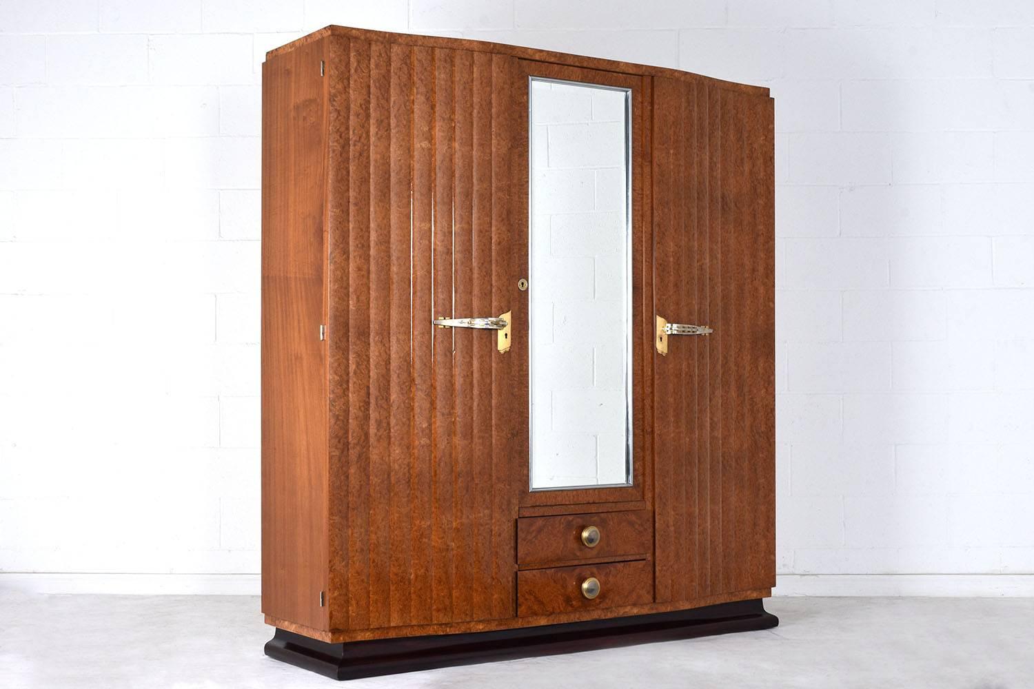 This 1930s French Art Deco-style armoire is covered in burled walnut veneers in the original light walnut color stain finish. There are two cabinet doors that have decorative wave details and brass door handles. The centre door features a mirror
