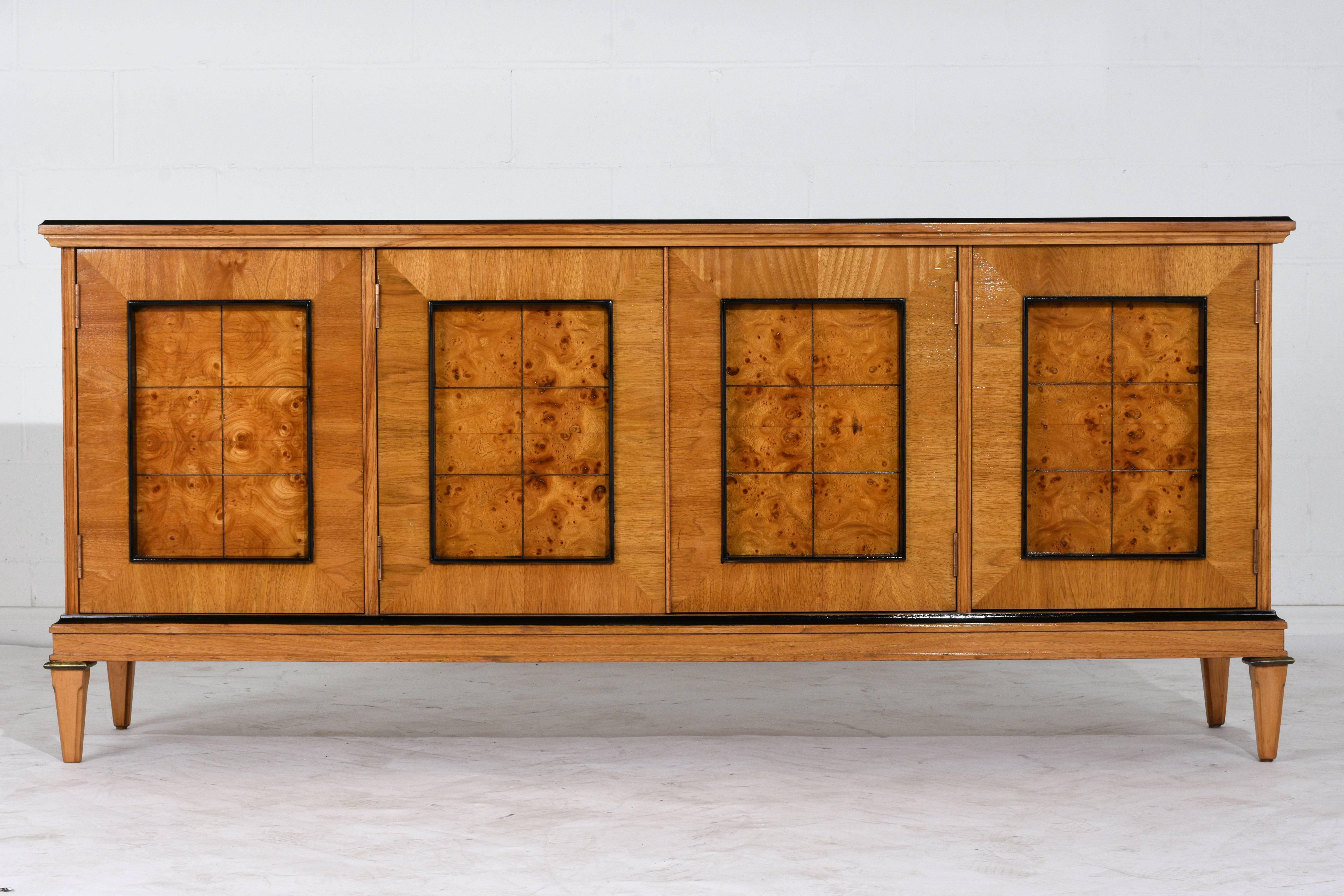 This 1950s Neoclassical-style buffet is made of wood covered in maple and walnut veneers finished in a light, golden color stain with a lacquered finish. The edges of the buffet are finished in a rich black color. The four cabinet doors have large