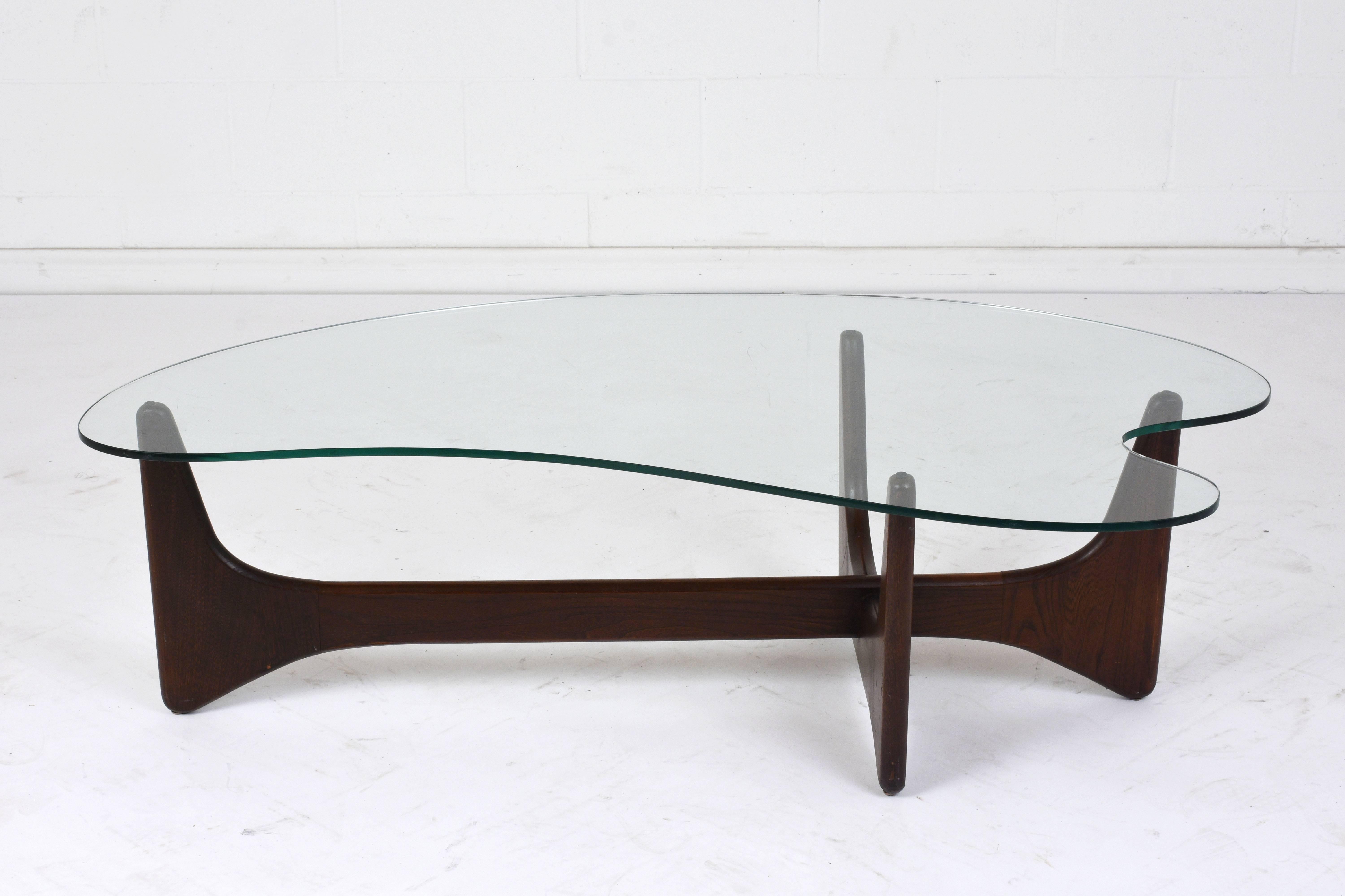 This 1970s Mid-Century Modern style coffee table is made in the style of Adrian Pearsall. The cross shaped walnut wood base features curved edges and is sturdy. The unique shaped glass top resembles an artist's palette. This coffee table is sturdy,