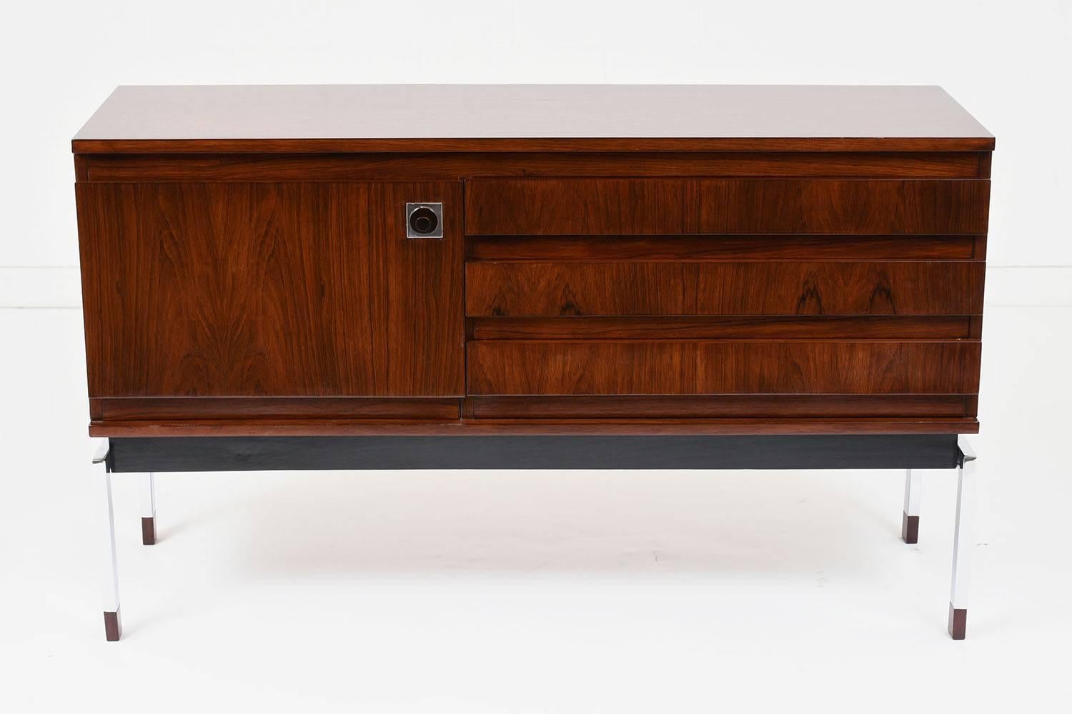 This 1960s Mid-Century Modern style credenza is made of rosewood stained in a rich rosewood color with a polished finish. The credenza has three drawers with carved finger pulls on the right and a cabinet with a knob pull on the left. Inside the