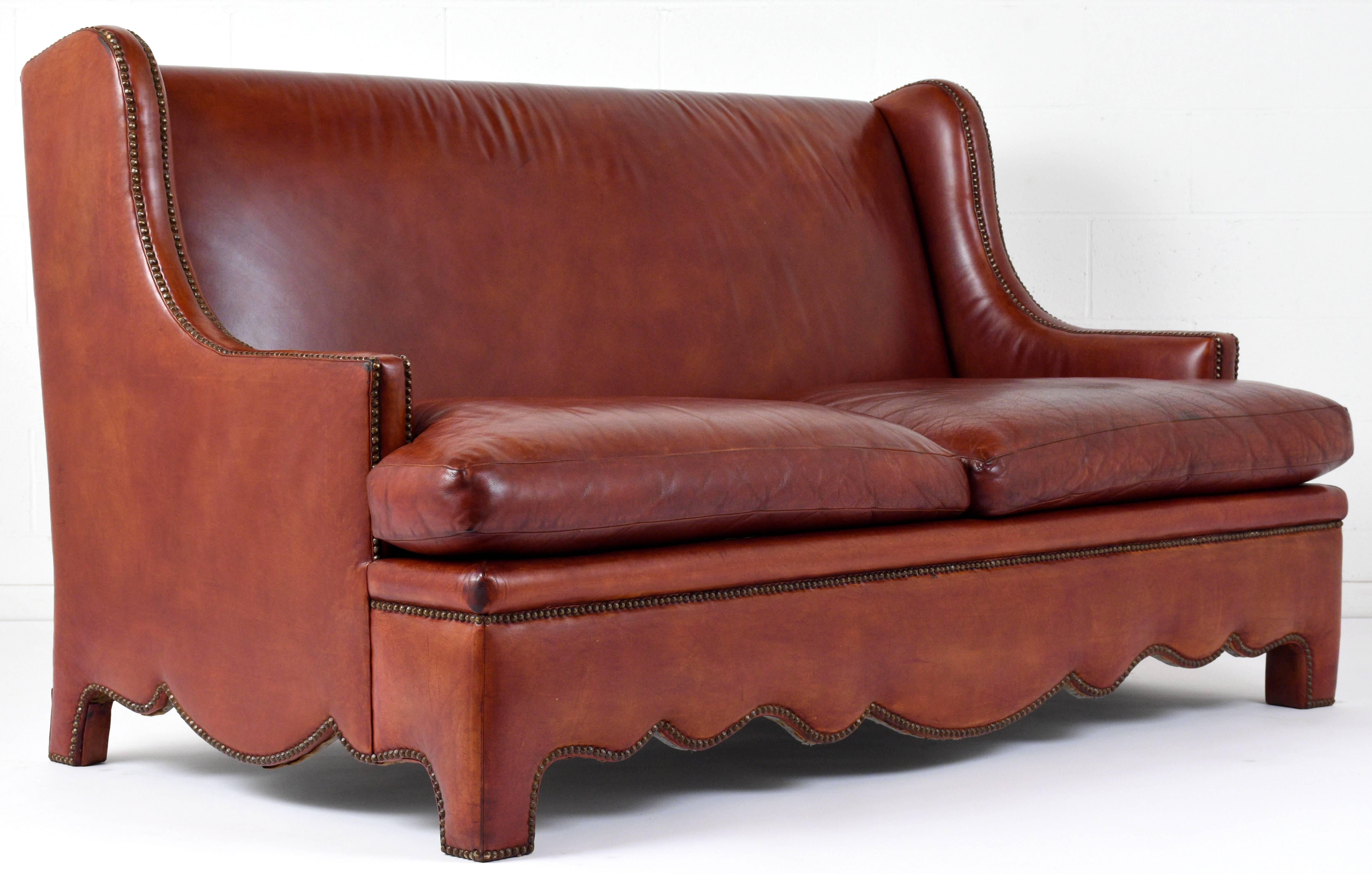 This 1970s vintage Regency-style sofa is fully upholstered in the original leather in a tan color. The high back seat with curved arms and the scalloped bottom edges have nailhead trim decoration. The seat has two loose cushions with comfortable