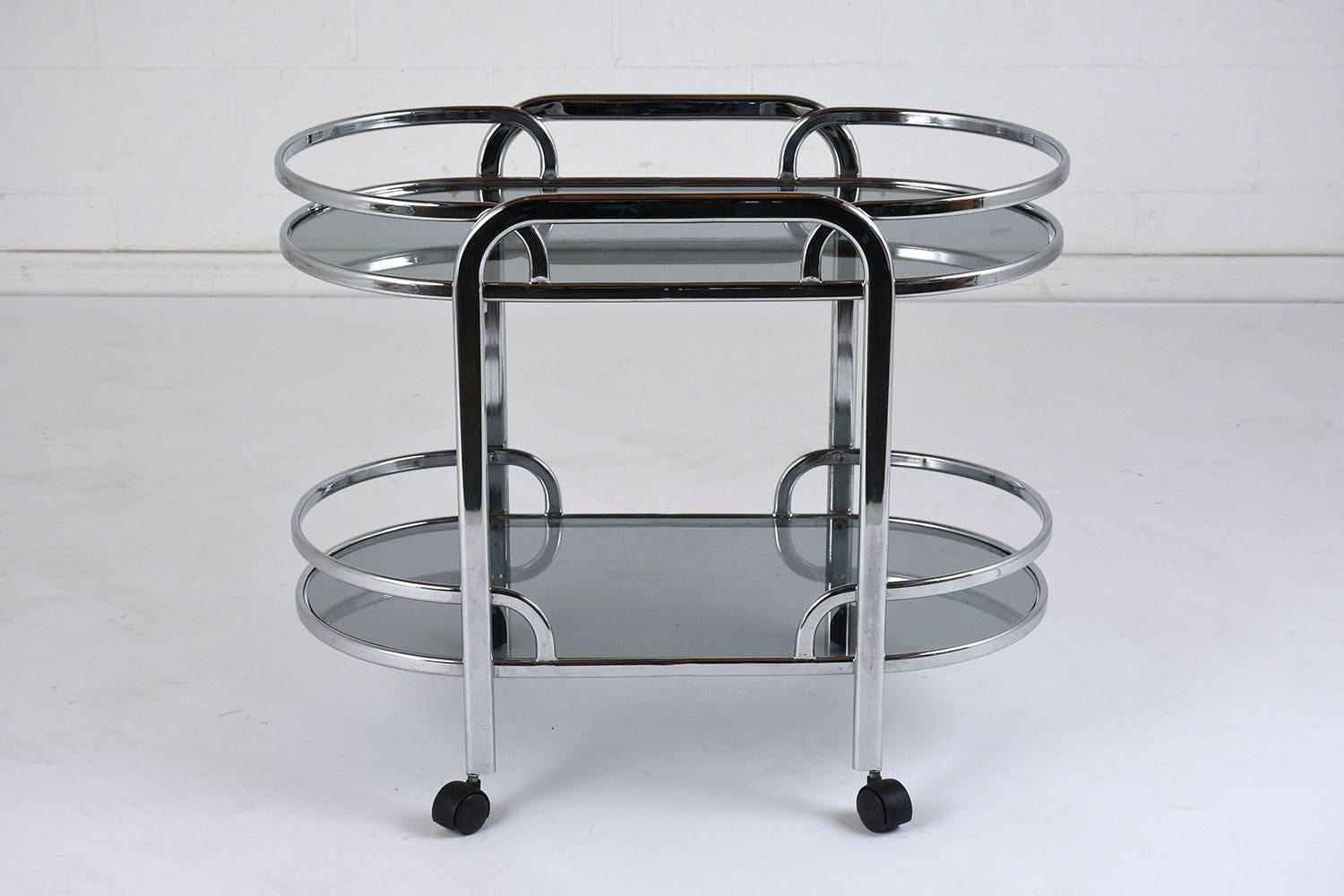 This 1960s Mid-Century Modern style bar cart features a chromed steel frame and smoked glass shelves. The top and bottom tier mimic each other with curved edges and flat rails. The handles on the sides of the cart lead down to the wheels. The caster