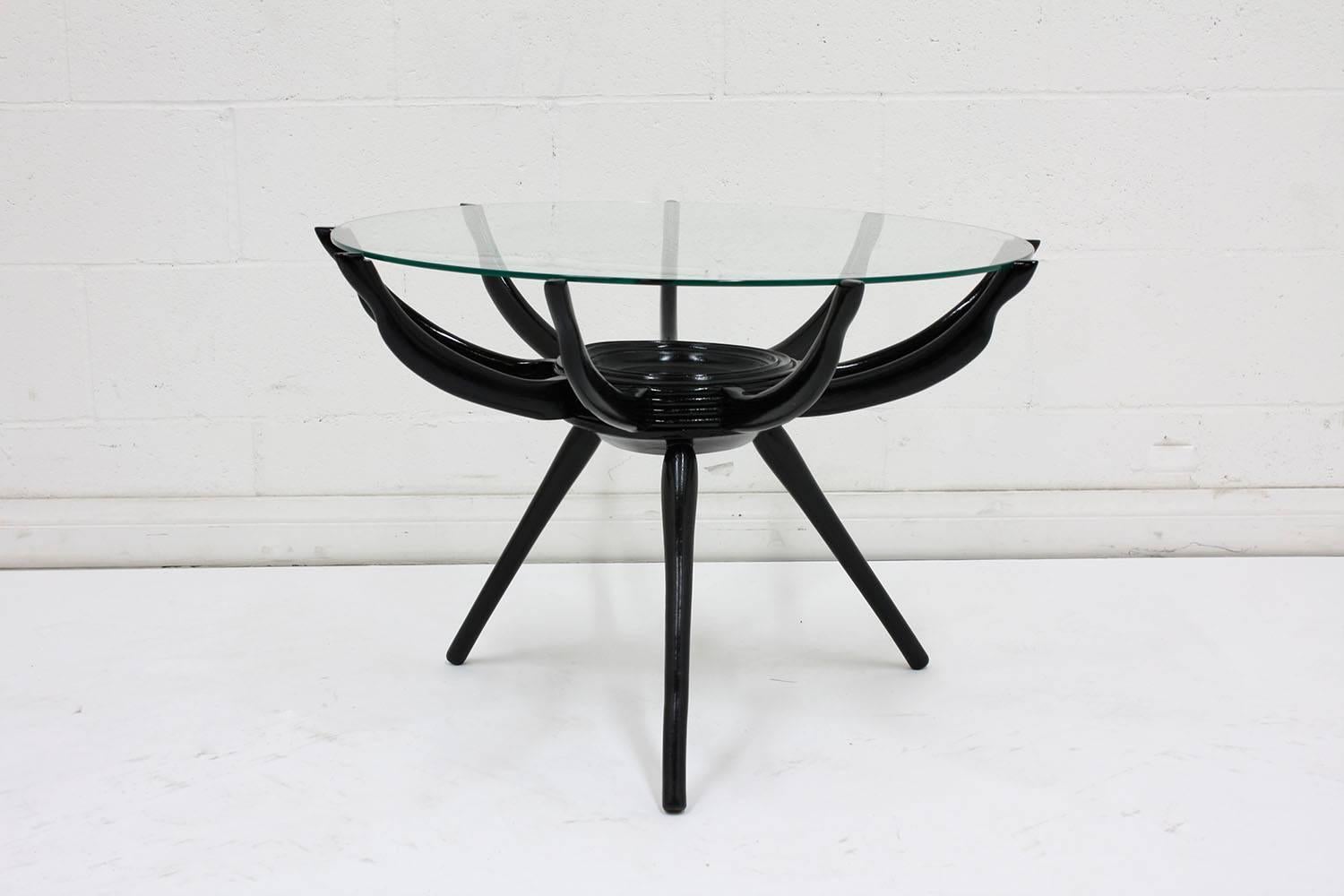 This 1960s Mid-Century Modern style coffee table features a wood base and a glass top. The pedestal wood base has an ebonized and lacquered finish. The base has a circular centre with nine arms curving from there to hold up the glass top and three