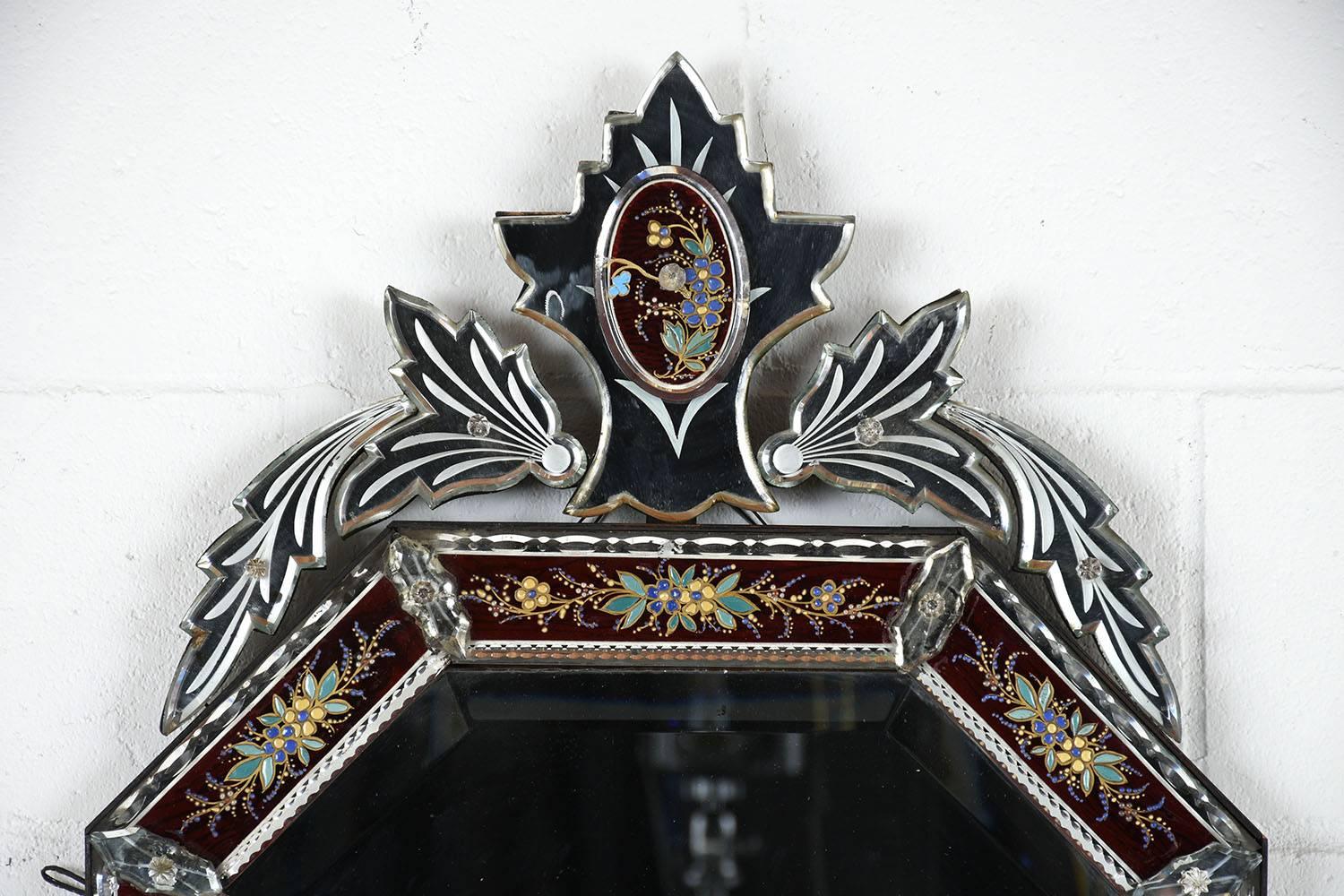 This 1890s Italian Venetian-style mirror features a Murano glass and mirror frame. The decorative frame has a leaf shaped crest with etched details and raised glass accents. Adorning the frame is raised enamel and gold color paint floral and leaf