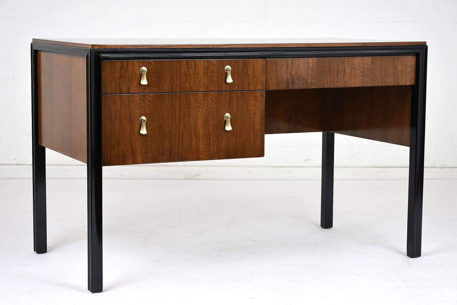 This 1960s Mid-Century Modern style desk has been completely restored and features walnut wood veneers stained in a rich walnut color with a lacquered finish and black details on the corners and legs. There are three drawers with brass drop drawer