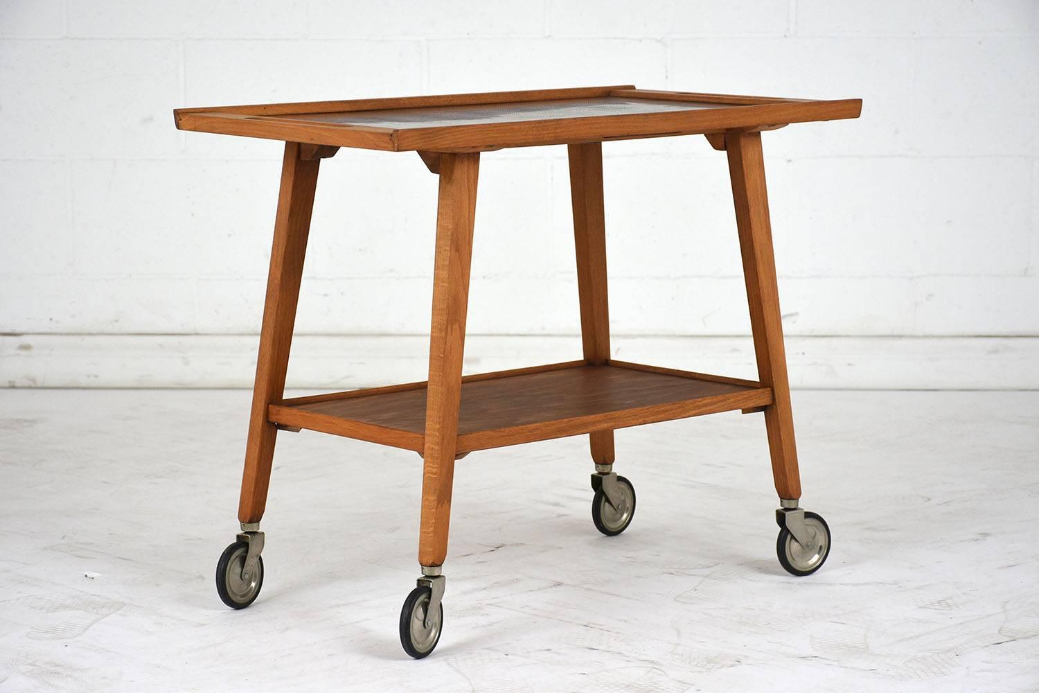 This 1970's Mid-Century Modern-style bar cart features a wood frame stained in a light walnut color. The top of the cart has an inlaid detail of feathers and handles on the side. Below is an open shelf. The bar cart is finished with tapered legs and