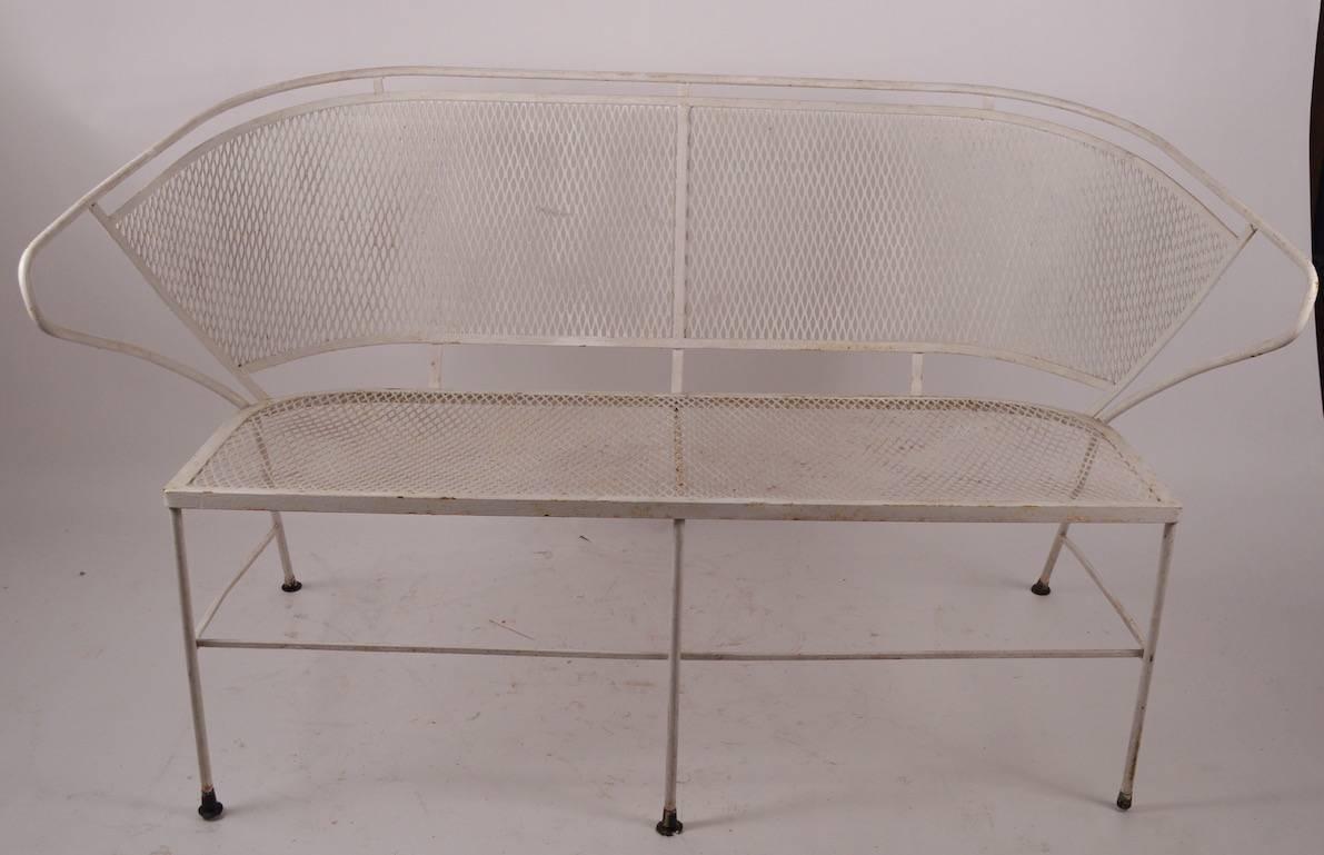 Interesting wrap around backrest metal mesh and wrought iron settee, attributed to Woodard Furniture Company. Mesh seat and back, wrought iron frame and legs, plastic glide feet (some replaced). Currently in old white paint finish, which shows wear