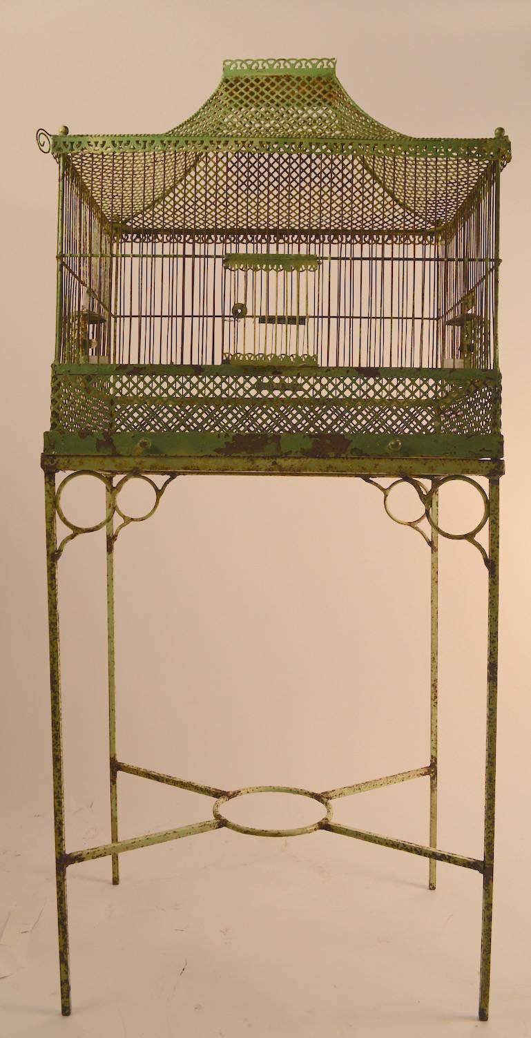 Metal bird cage on original stand, in green paint finish (shows wear) the cage has a slide out tray, two feeding stations, and a front and rear door. The rear door opening is missing a vertical bar on each side - this does not affect its function.