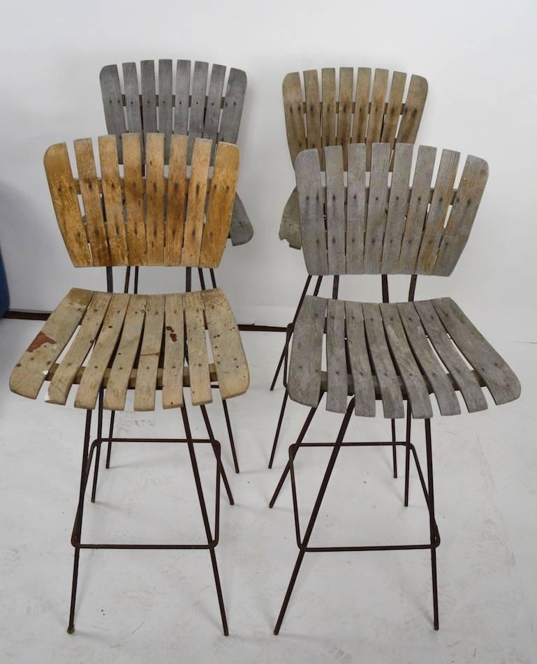Four swivel stools designed by Arthur Umanoff, in weathered finish. These stools are on squared iron bases (show rust), they are missing the foot cap glides. 
Three of the four are weathered grey, while one retains more of the original stained