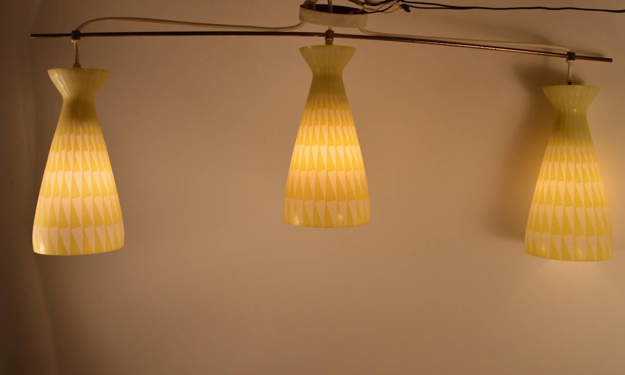 Interprétation moderniste d'un  forme classique de la lampe de billard. Trois abat-jour en verre de forme cintrée, avec un fond blanc et un décor jaune, sont suspendus à une fine tige en laiton, elle-même suspendue en son milieu à la verrière du