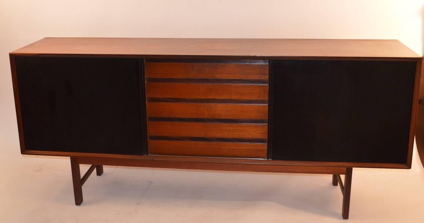 Danish modern teak credenza, with sliding black doors and five drawers in the middle section. The door open to reveal storage with an adjustable shelf.