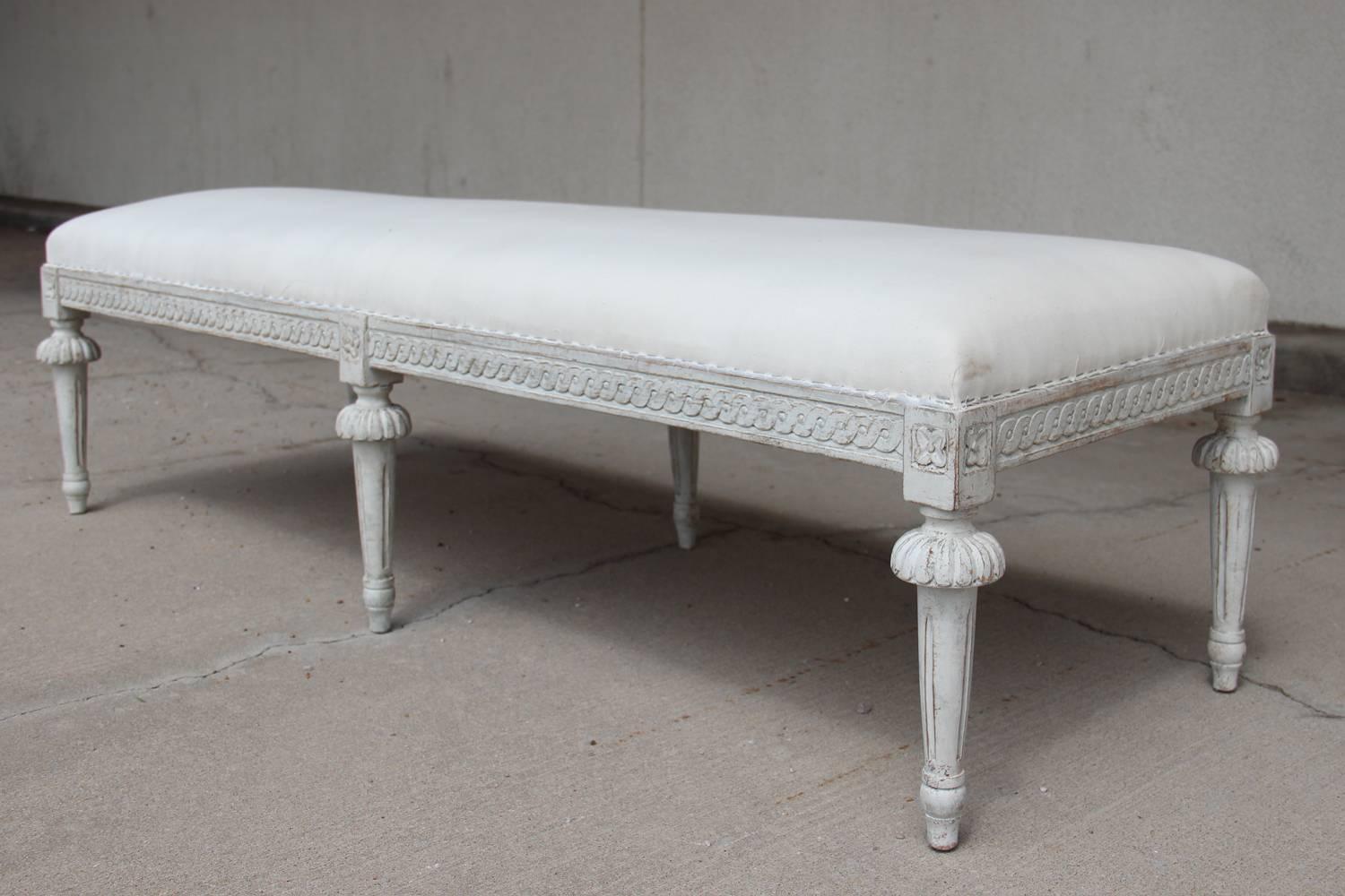 A richly carved 19th century Swedish painted long bench from the Gustavian period with carved guilloche pattern on the seat frame and carved rosettes above fluted, round caps, ending in tapered and fluted legs.  Chalky Gustavian white paint finish.