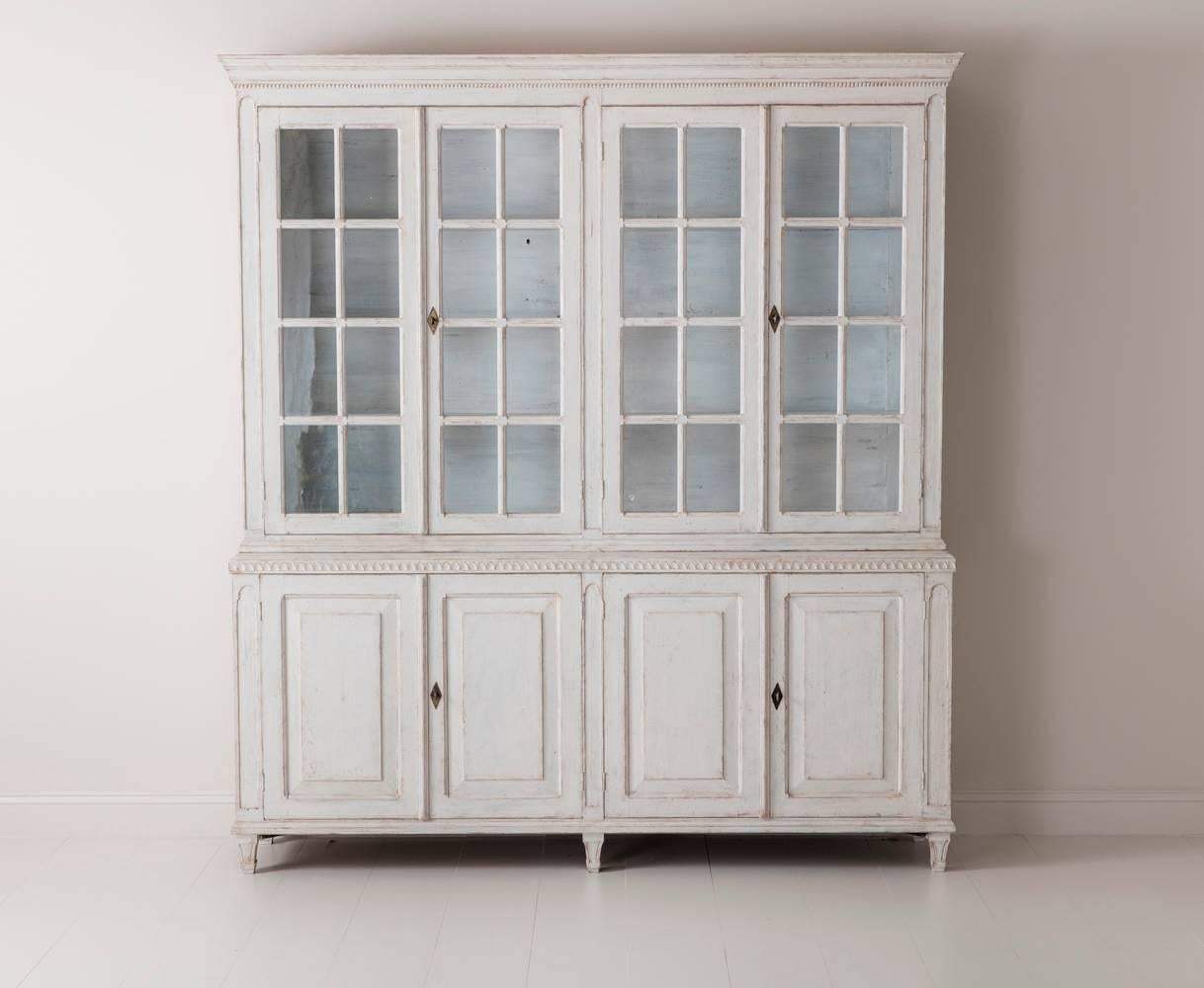 A beautiful Swedish Gustavian style four-door painted vitrine cabinet from the 19th century. This bookcase is made in two parts. The upper section has three fixed shelves behind original glass doors. 
The shelf depth is 10 3/4 inches at the bottom