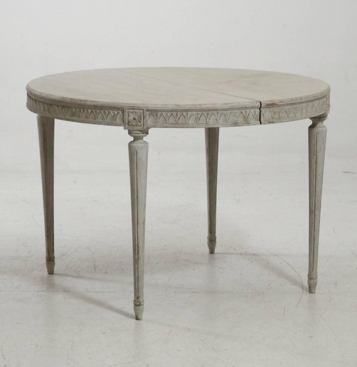 A charming Swedish three-leaf oval dining table in the Gustavian style.  The patina is a blend of light gray and aged white.  There is beautiful carved egg and dart molding around the apron with a carved floral motif above each round, tapered, and
