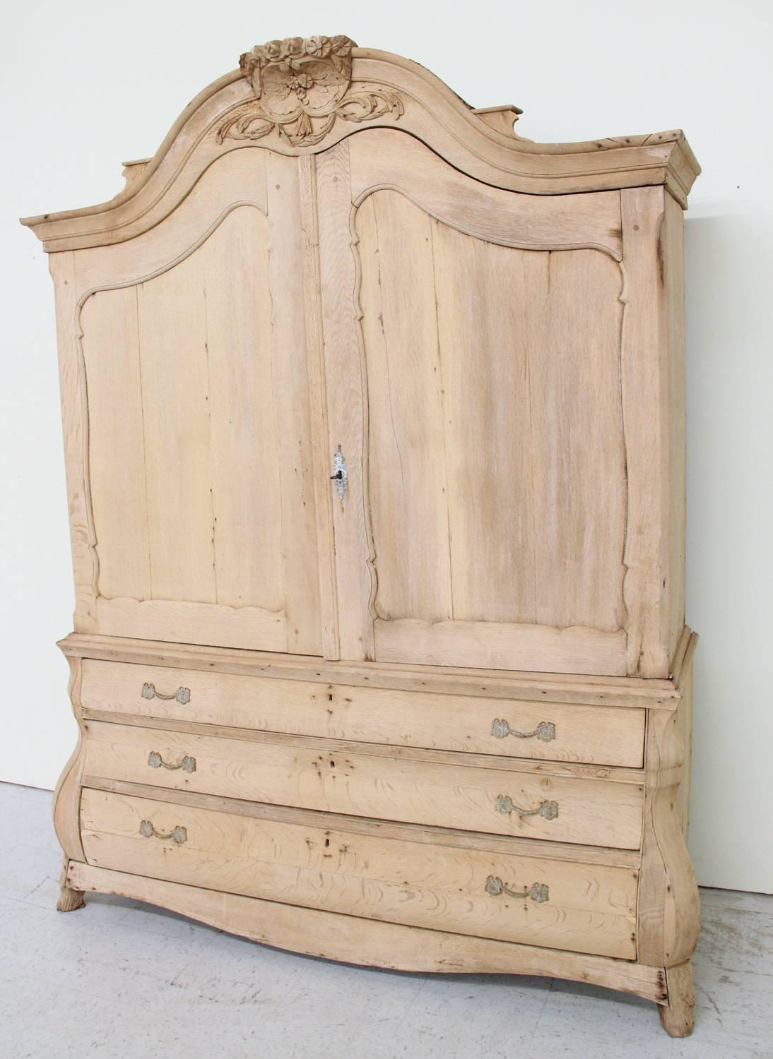 A magnificent hand-carved, two-part cabinet with brass hardware, beautifully aged. This 19th century Dutch Louis XV style bleached oak armoire has a richly carved cornice and three-shaped lower drawers, resting on carved paw feet. The gray painted