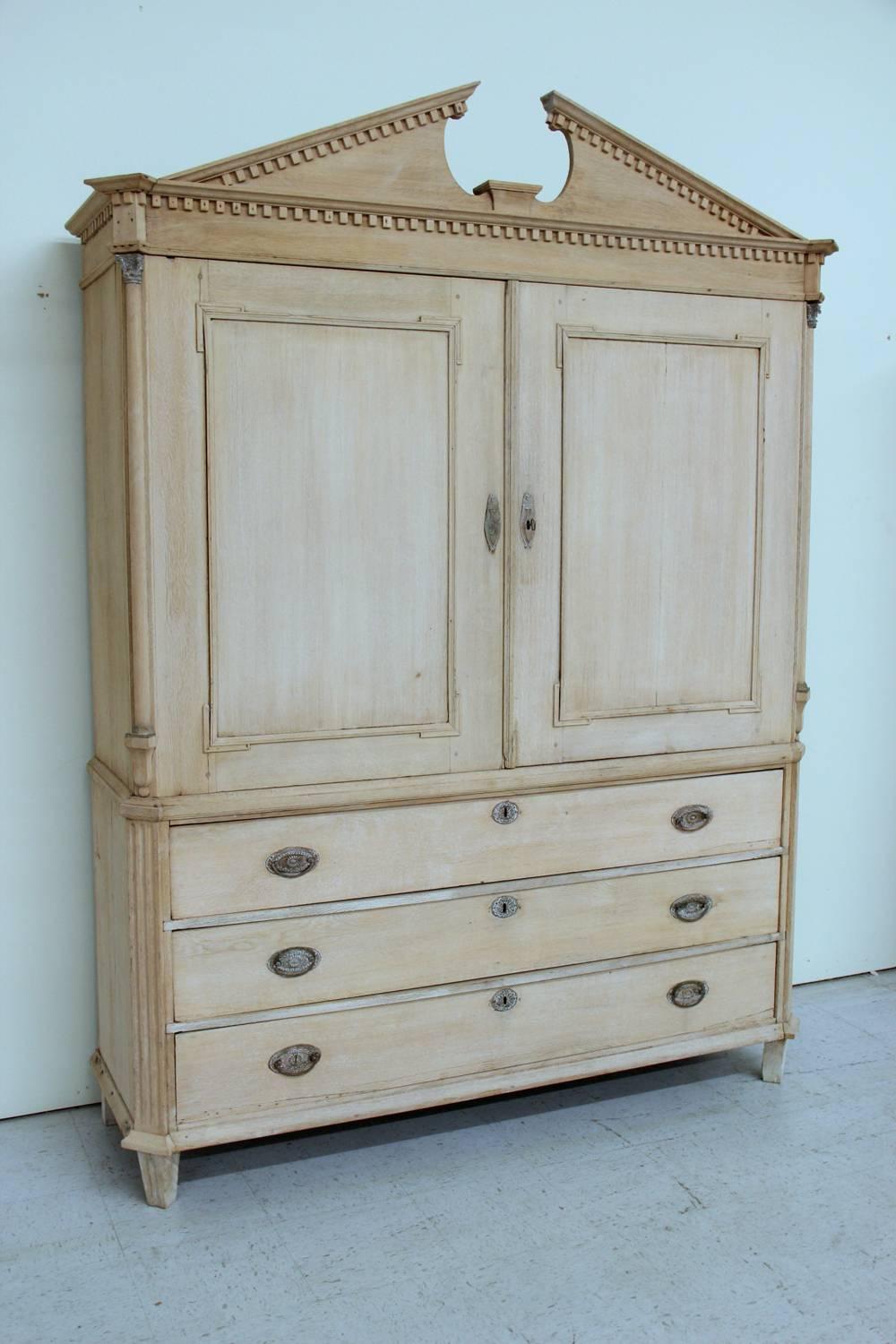 19th century Dutch neoclassical, two-part armoire in bleached oak with brass hardware and three large lower drawers. This cabinet has a pediment framed by dentil molding and half-round columns with original brass mounts. The grey painted interior