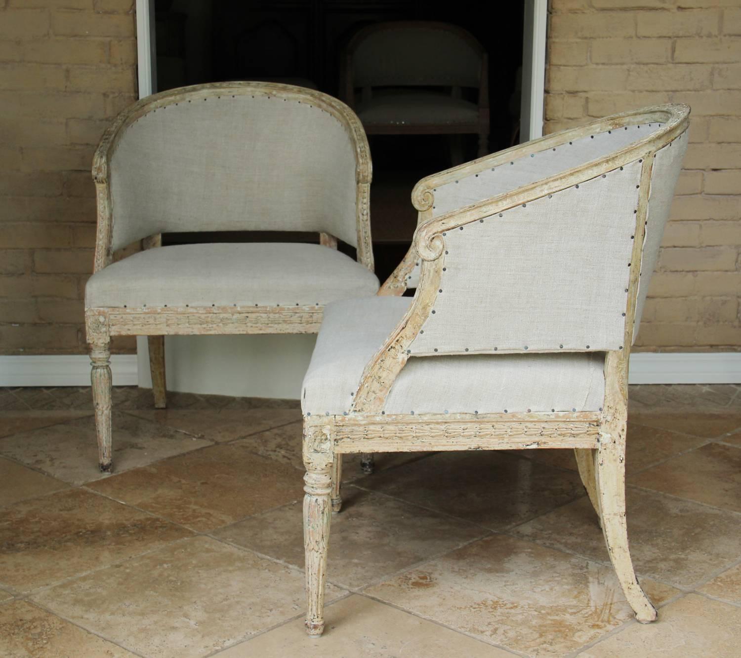 A pair of Swedish Gustavian period armchairs in original paint with barrel backs. These comfortable chairs have been newly upholstered in antique French linen. There is laurel leaf carved molding along the back frame ending in subtle scroll arms.