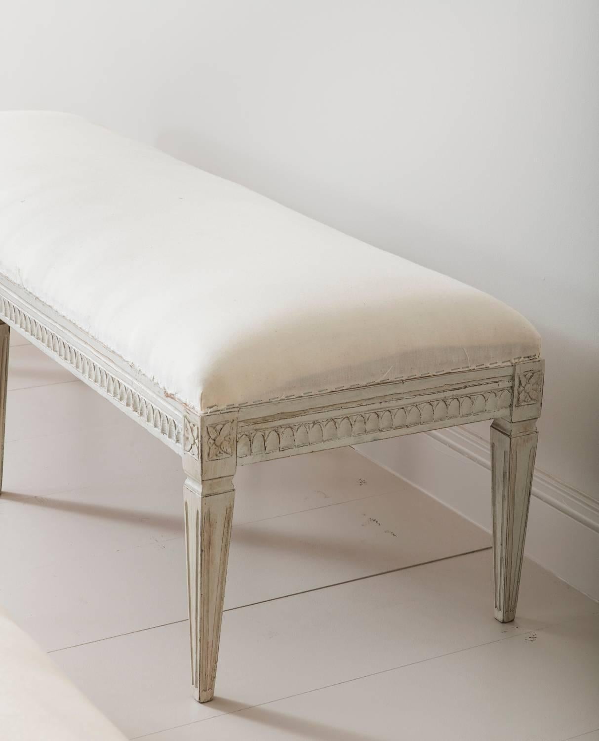 This is a richly carved 19th century Swedish Gustavian period painted long bench. There is carved egg and dart detail on the seat frame and carved rosettes above tapered and fluted legs. Chalky Gustavian gray-white paint finish.

The Gustavian