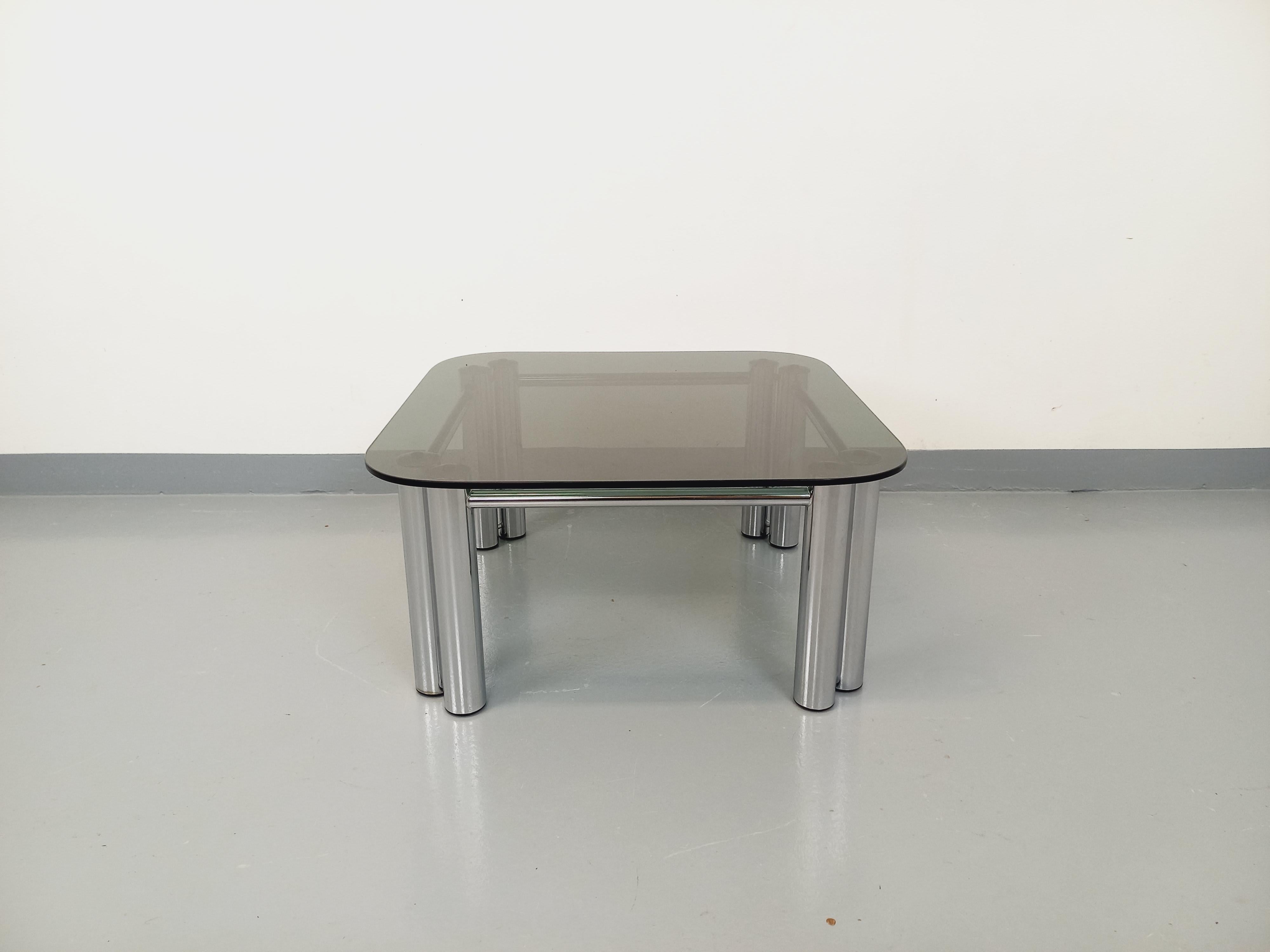 Table basse carrée vintage des années 70, très beau design italien style Space Age, plateau carré aux angles arrondis en verre fumé, pieds doubles cylindriques et structure tubulaire design en métal chromé. Très bon état général avec quelques signes
