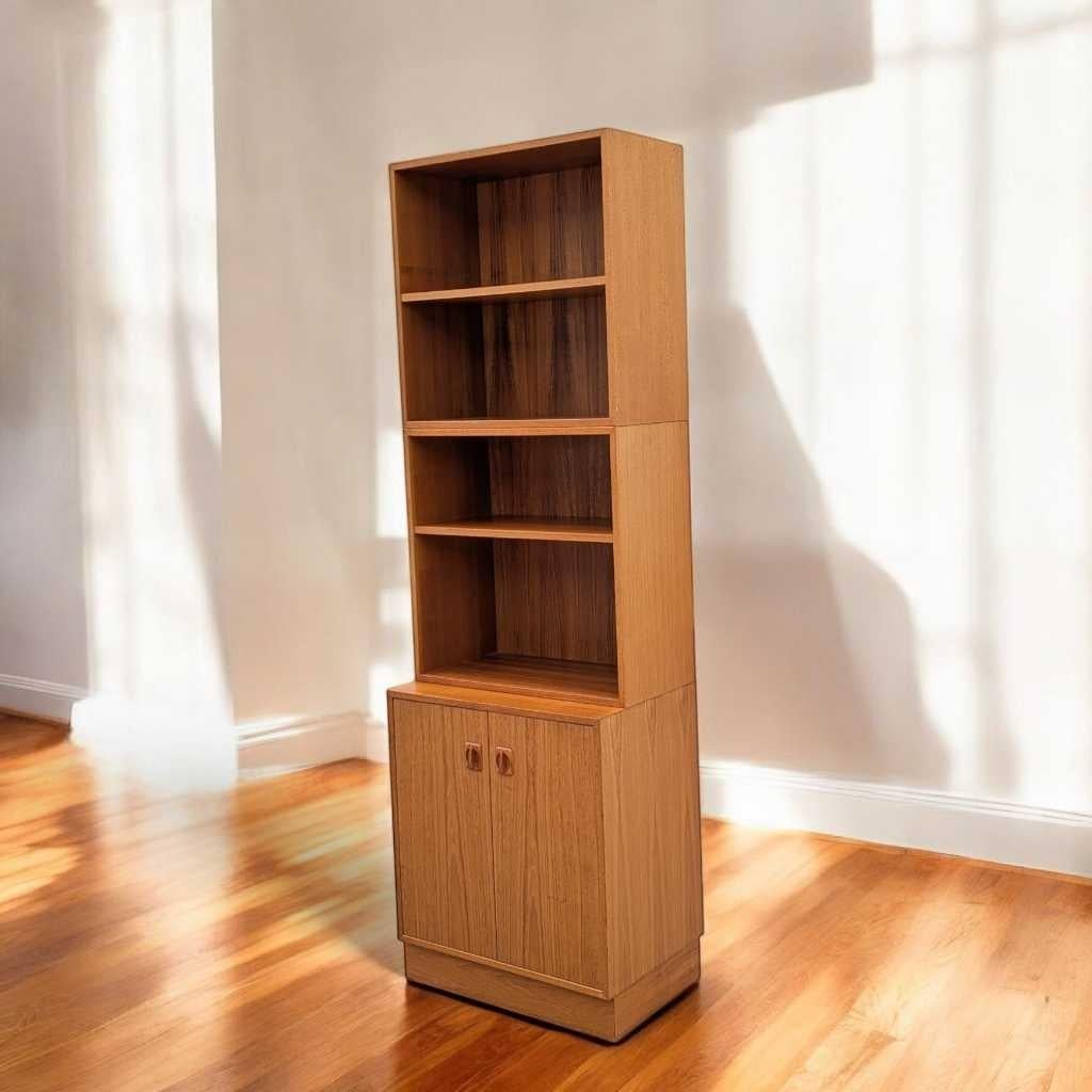 A distinctive bookcase in the mid-century modern Scandinavian tradition, crafted from richly grained teak veneer and showcasing a sleek, upright profile with open shelving above and a closed storage cabinet below. This modular piece blends warm,