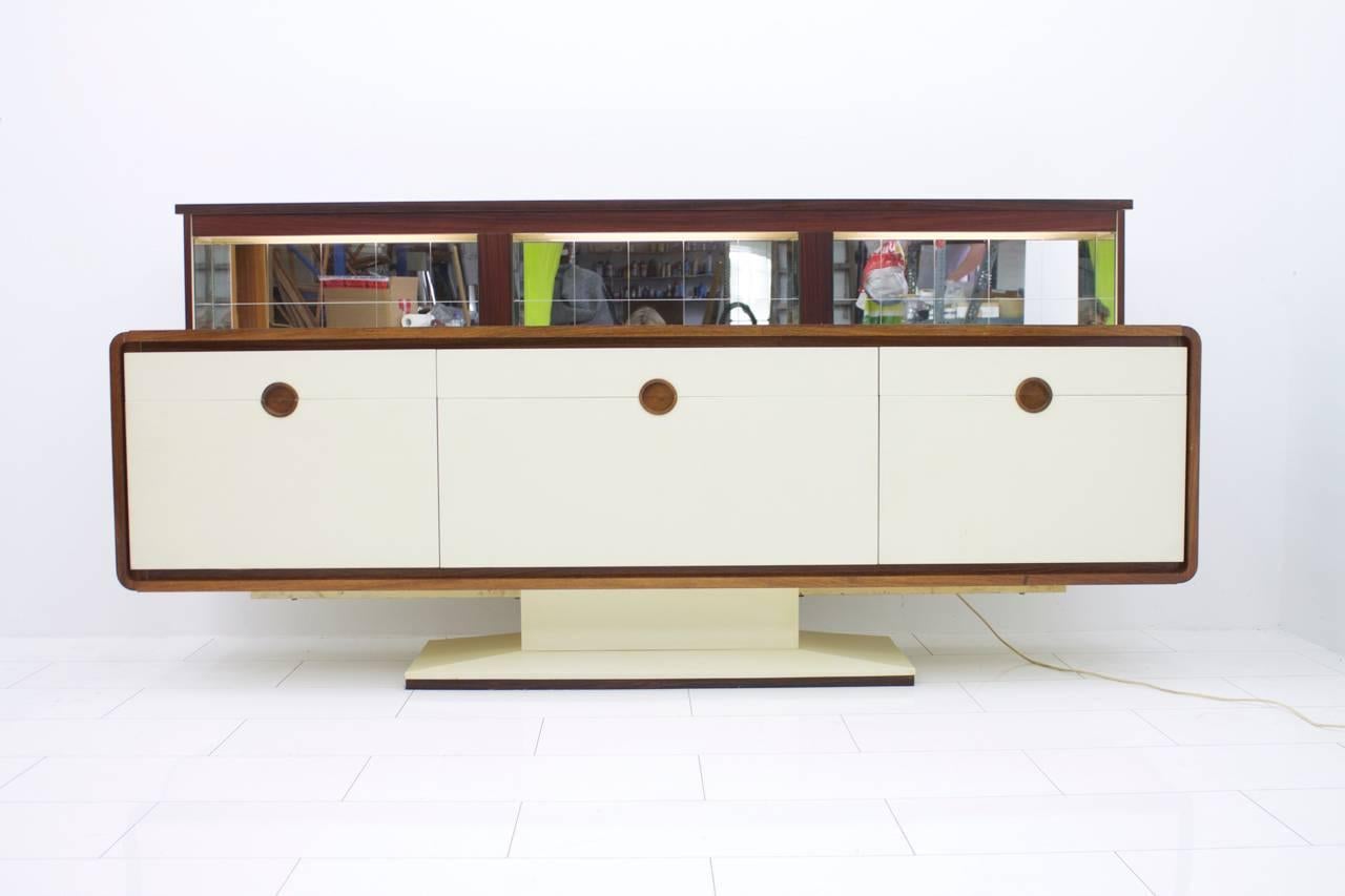 A special sideboard with an electric bar and mirror, Germany, 1970s.
By pressing a button moves up from the rear part a illuminated mirror Bar.
The sides and the surface is veneered with wood. The Front and the Foot are coated with