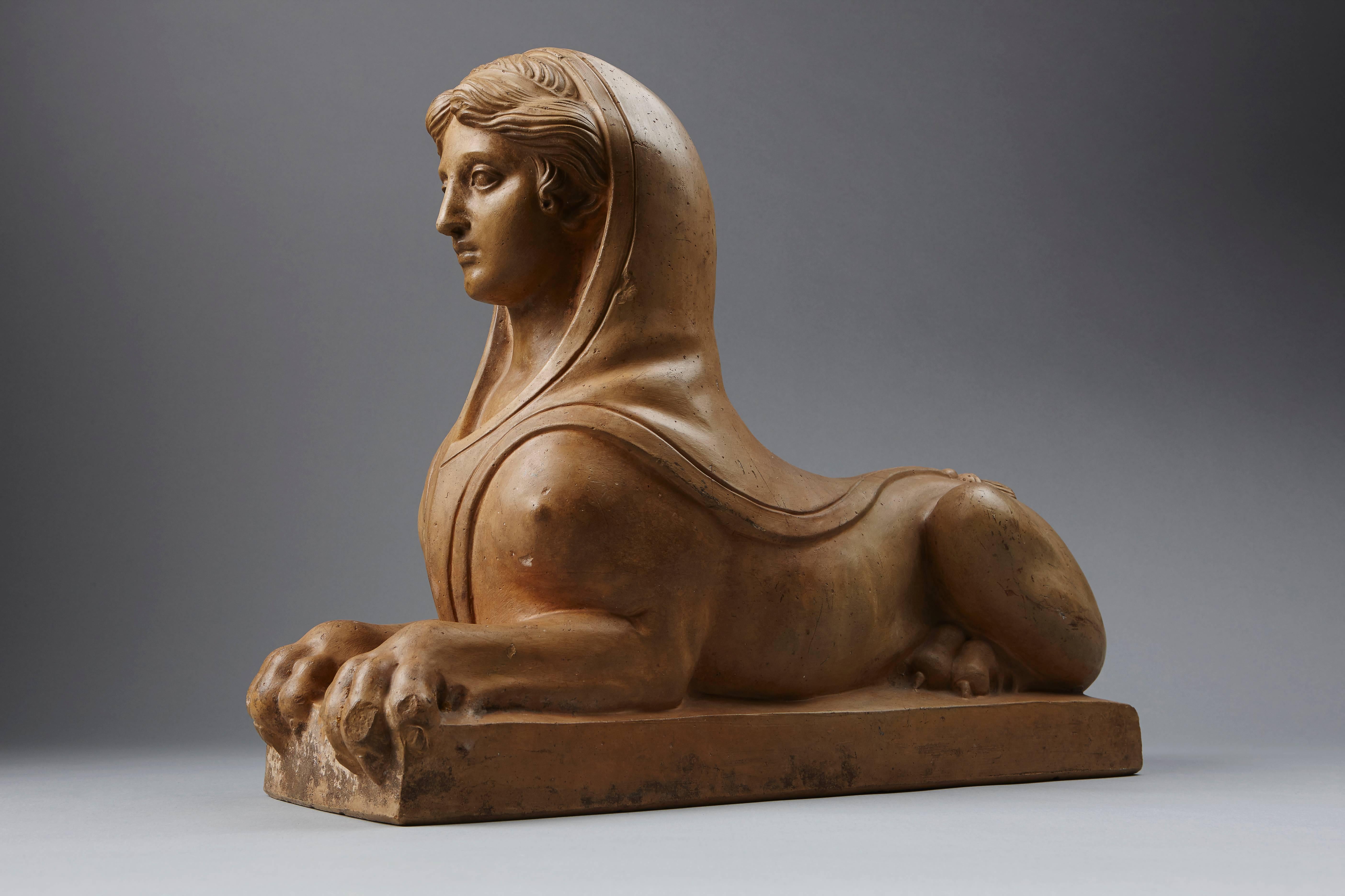 A very decorative early 19th century empire Prussian terracotta figure of a reclining sphinx on a rectangular plinth, with serene female face wearing a loose hood draped around the head and chest, extending to a tassel on the rump.

Probably the