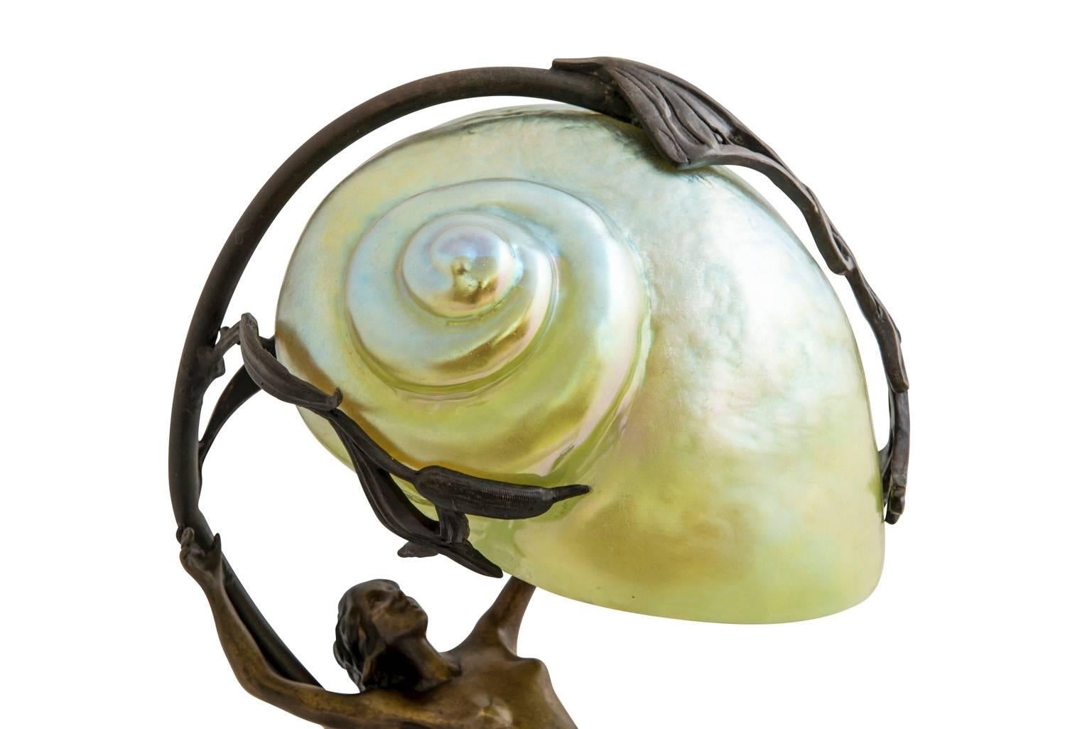 This lamp was produced after a design by Gustav Gurschner in the 1920s. There is a picture in the archives with the number GG-1900041 with different foot. A mermaid riding on a wave, carrying an iridescent glass shell above its head is a typical