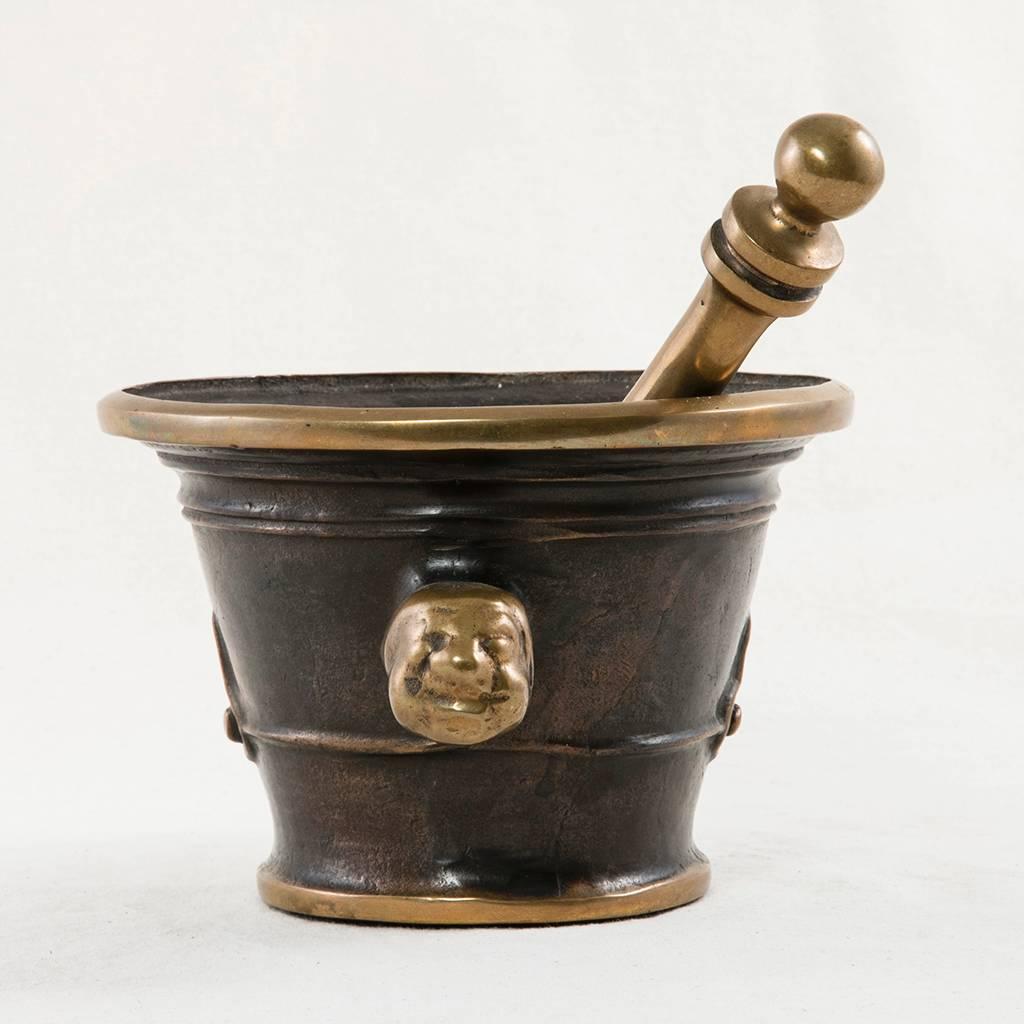 This fantastic bronze mortar and pestle features unusual protruding faces as handles. A heavy and substantial piece probably used by a French Pharmacy, this vessel has a deep dark patina contrasting beautifully with its polished smooth edges. The