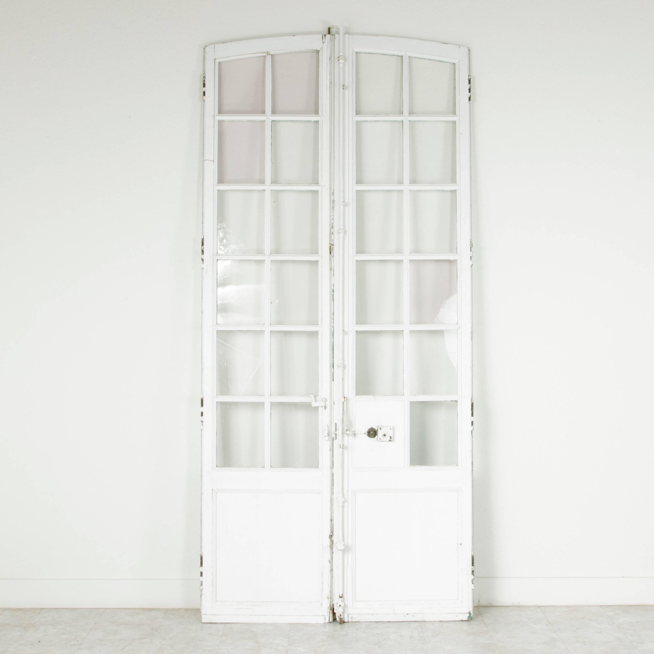 This stunning pair of doors were originally designed to hang in a French chateau, circa 1780. Each door is constructed of solid hand pegged wood painted in white and features 12 panes of original handblown glass. Their arched top stands at almost 10