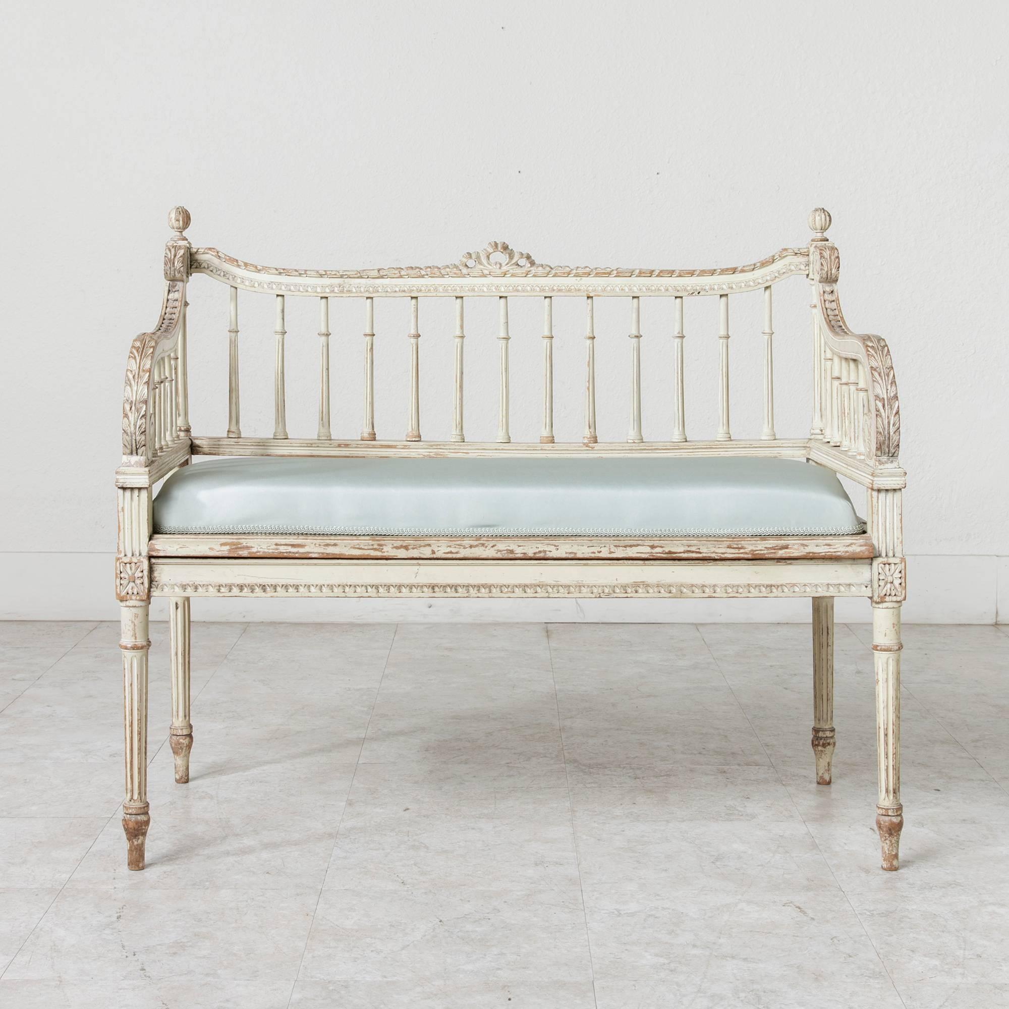 This hand-carved and painted Louis XVI style banquette, with newly upholstered in taffeta satin, features scrolled arms appointed with carved acanthus leaves. A colonnade of fluted columns embraces its sides and back and a bow and ribbon motif