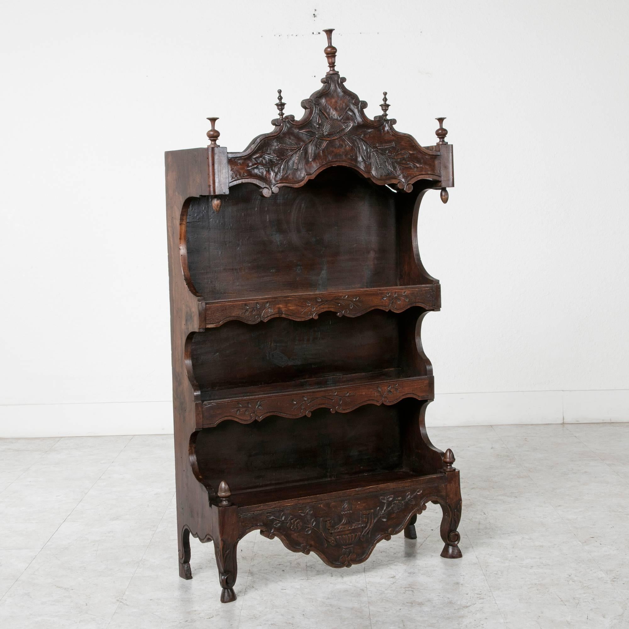 This early 20th century hand-carved walnut estagnie or etagere features carvings depicting a garden hat, garden tools, and shocks of wheat. From Provence in Southern France, its lower apron features the regional symbol of the Provençal soup tureen.